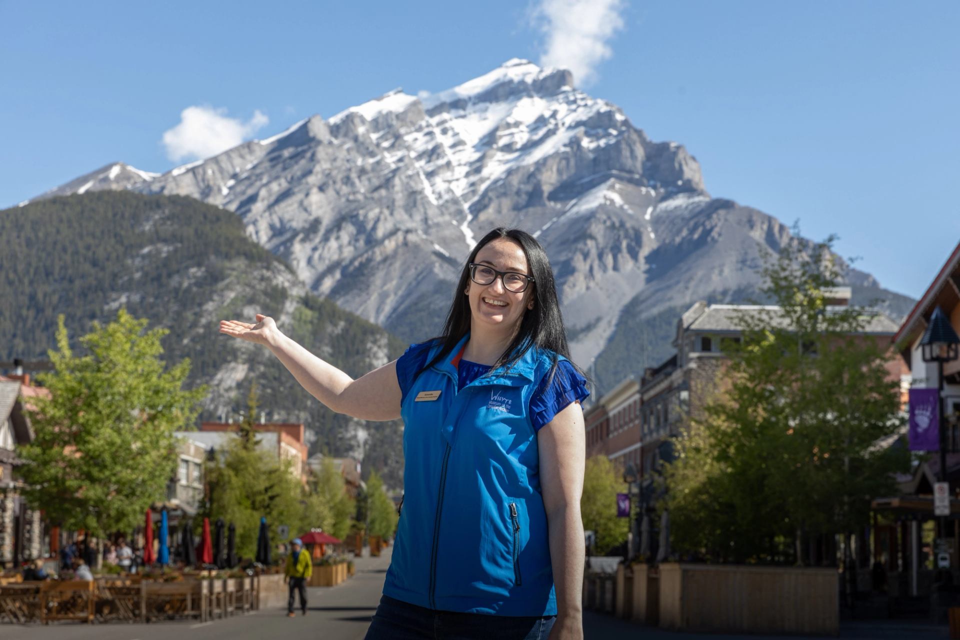 Person in blue vest gestures toward snow-capped mountain above town street.
