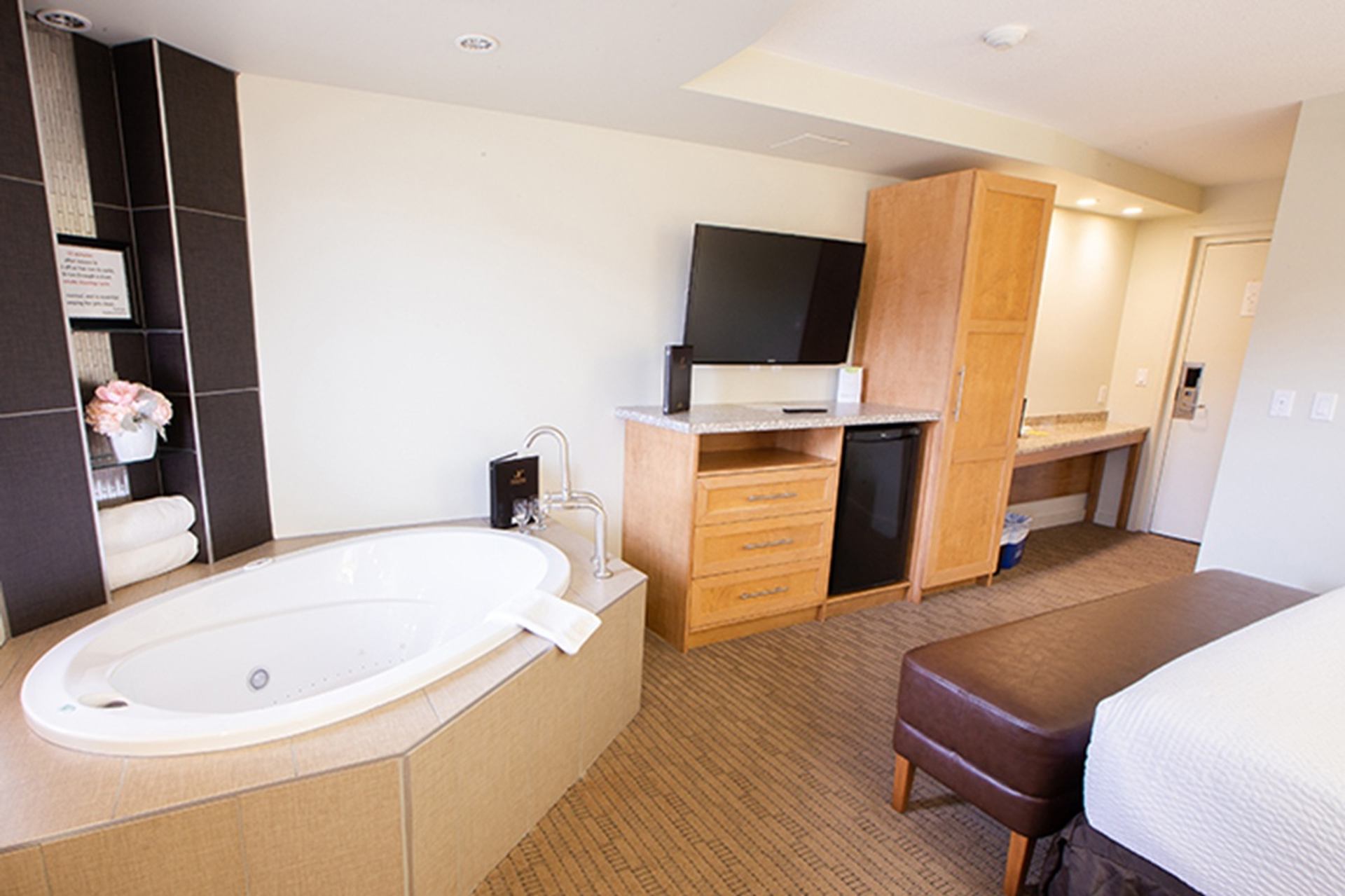 A guest room features a jetted bathtub beside a TV, mini fridge, and wooden cabinetry. A desk and luggage space extend toward the entry, creating a functional and comfortable layout.