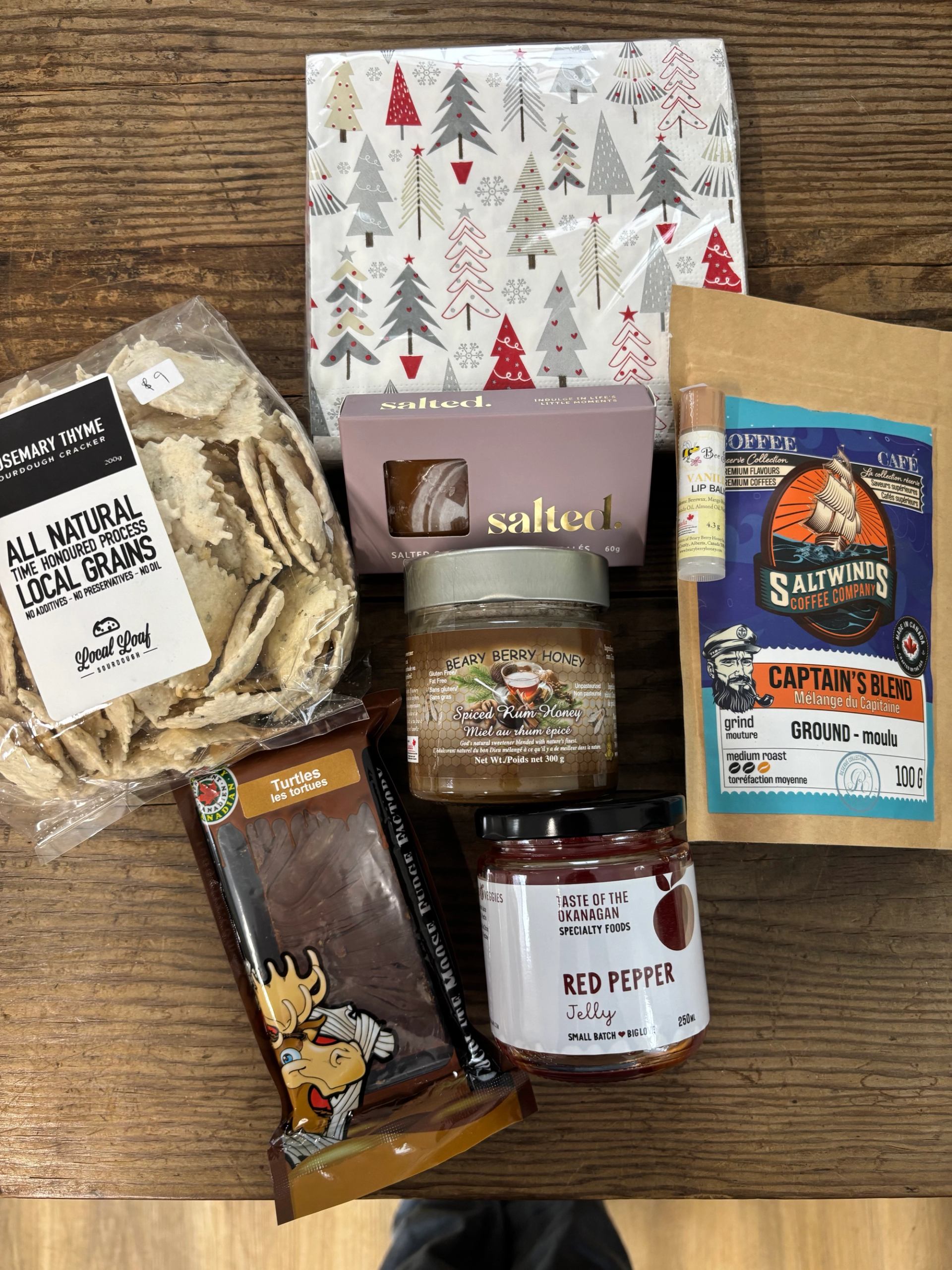 A collection of gourmet food products including crackers, honey, jelly, coffee, chocolates, and lip balm, arranged with Christmas napkins on a wooden table.