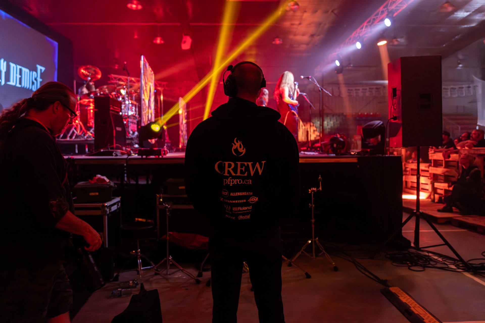 Decimate Metalfest crew member stands facing the stage with headset as band performs under red and yellow lights.
