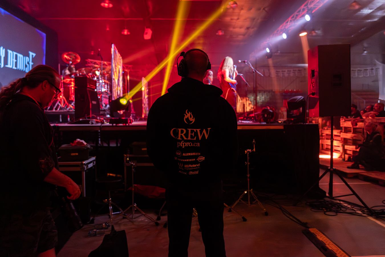 Decimate Metalfest crew member stands facing the stage with headset as band performs under red and yellow lights.