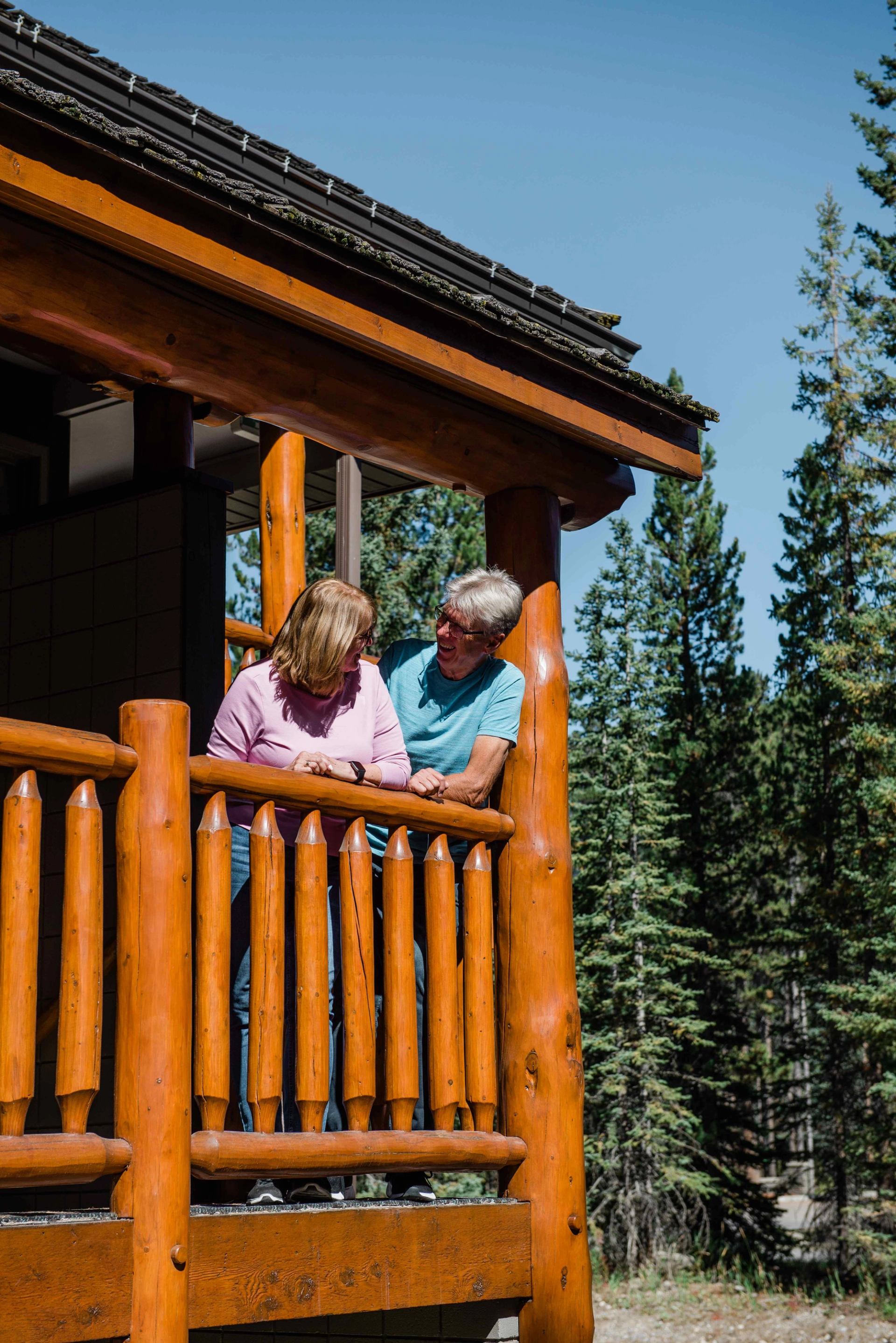 Two people on a log cabin porch at Mountaineer Lodge with trees and blue sky in the background.