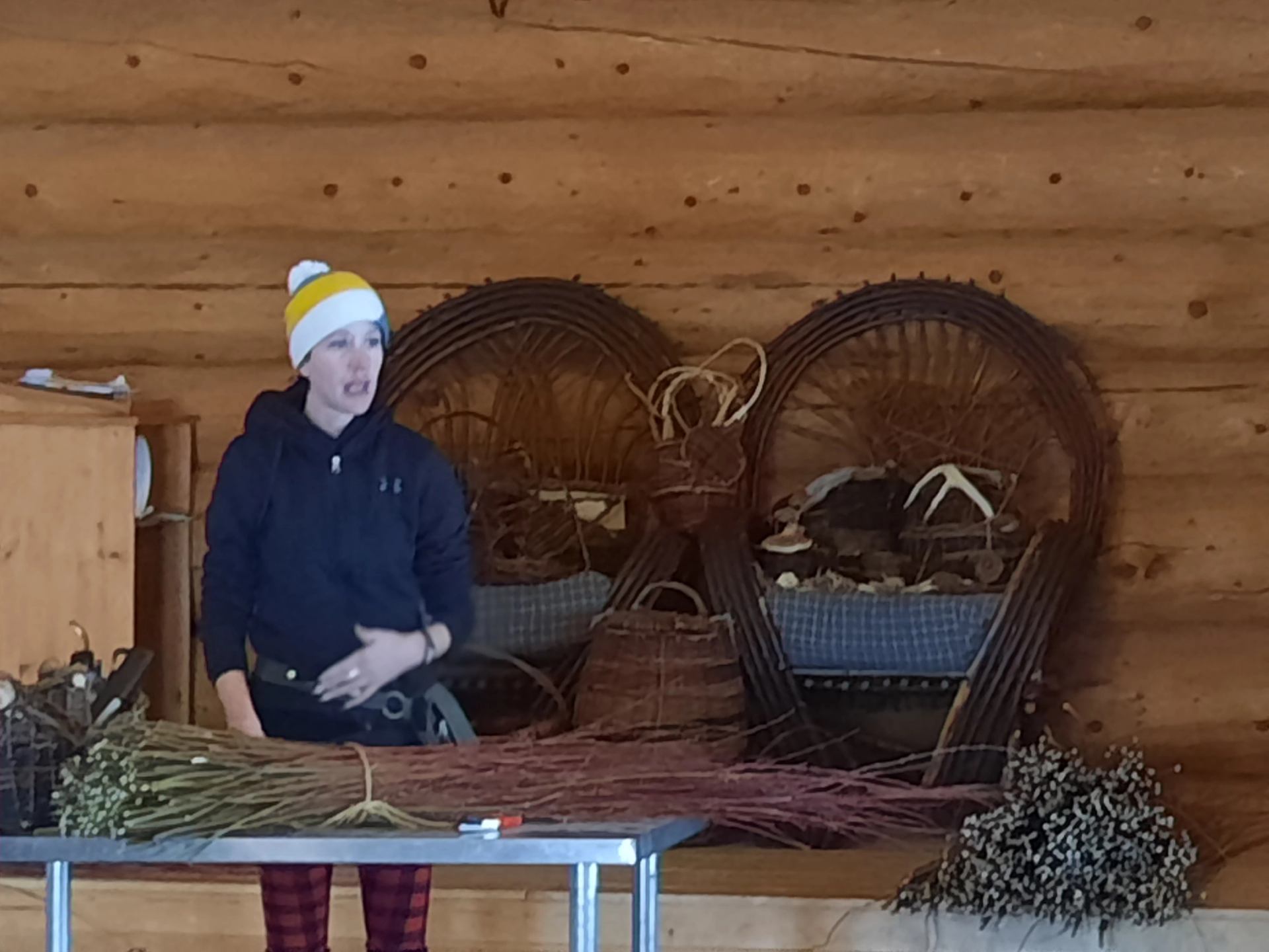 Person in toque stands at table with willow branches in rustic log cabin.
