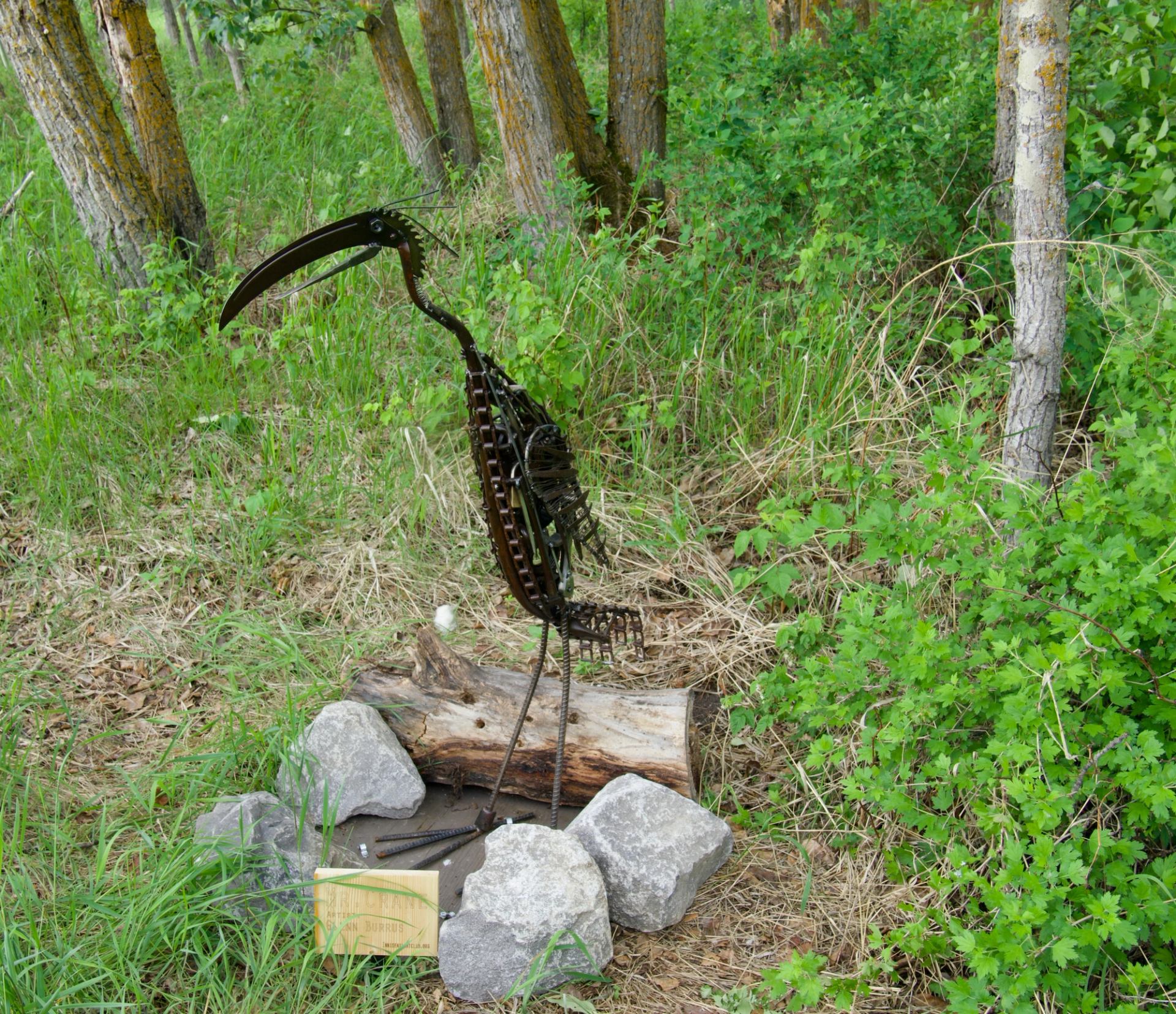 Metal bird sculpture stands by rocks and log among tall green grass.
