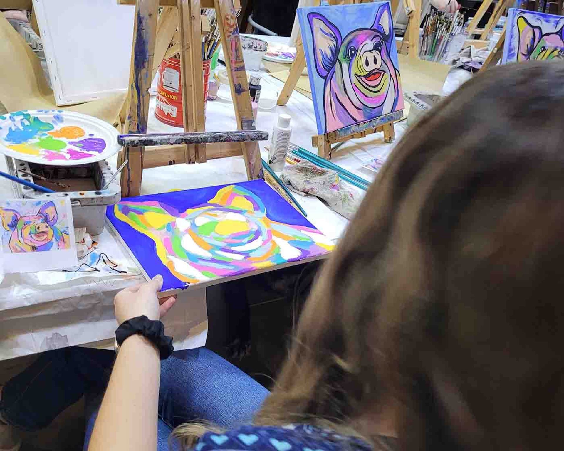 Painter holds a colourful pig portrait beside an easel displaying another pig painting.