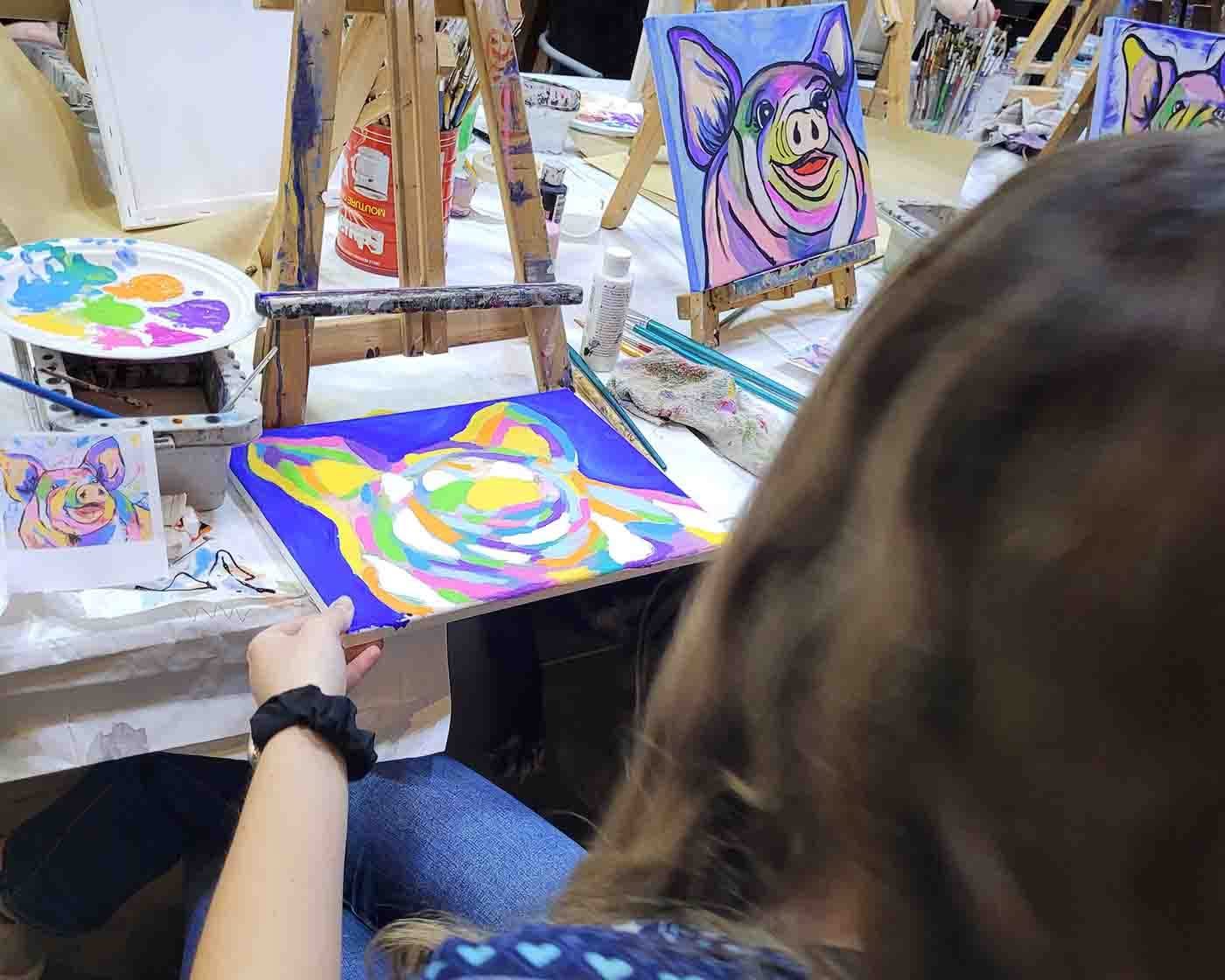 Painter holds a colourful pig portrait beside an easel displaying another pig painting.