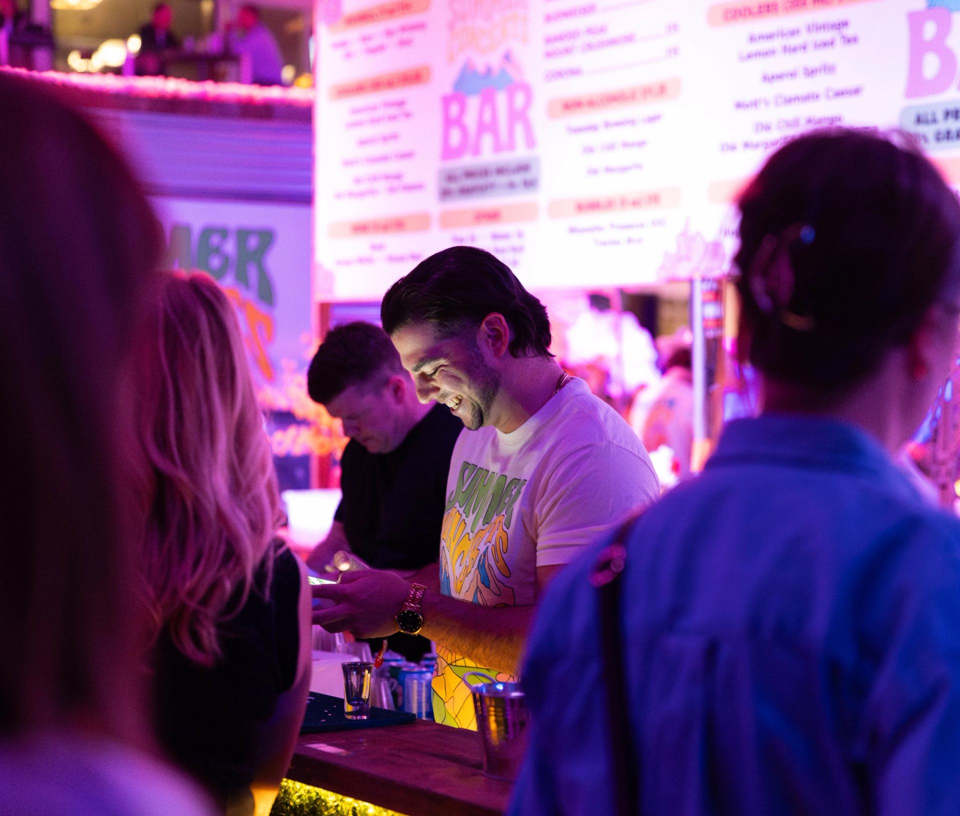 A smiling man stands at a bar counter, bathed in pink and blue neon light, with a drink menu board and other patrons in the background.
