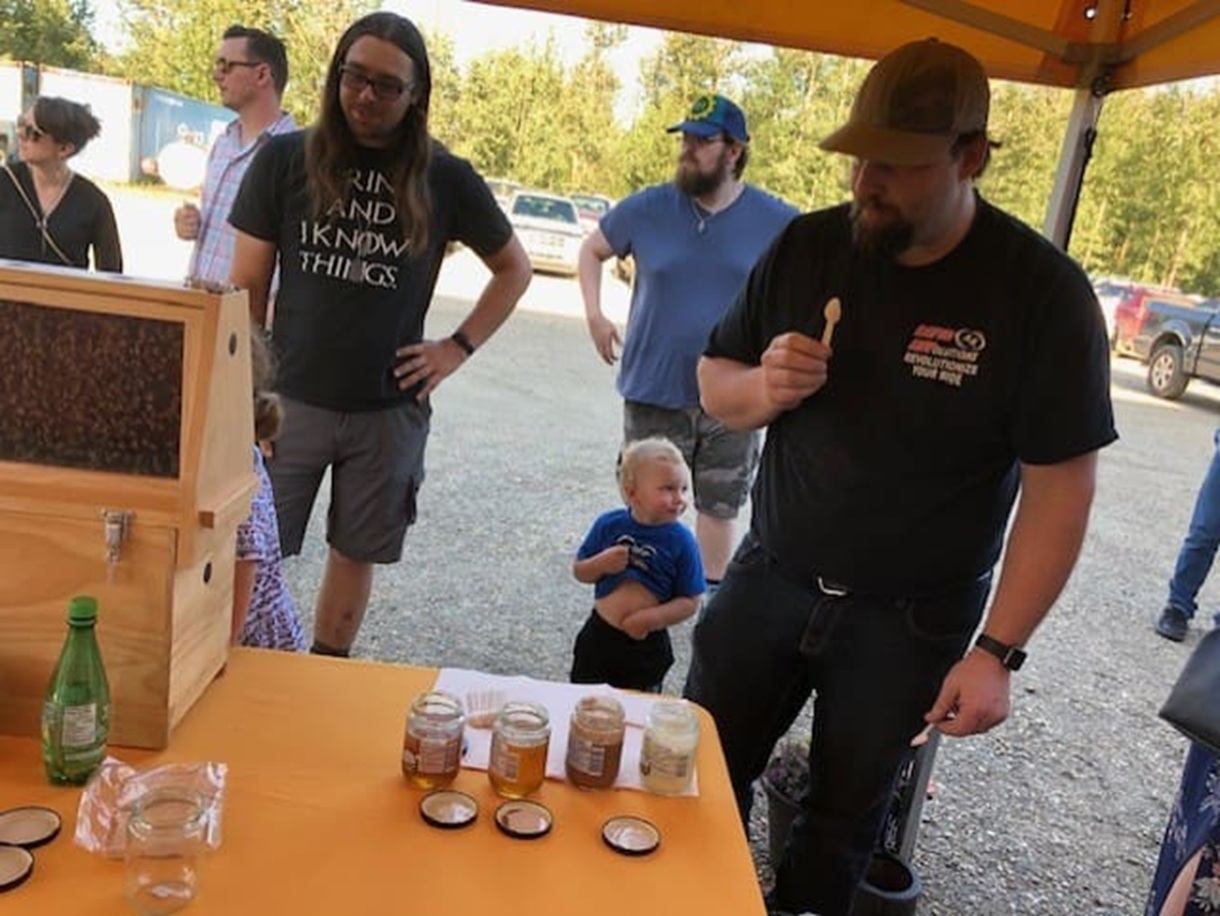 People sample honey at an outdoor market booth with jars and a bee display.