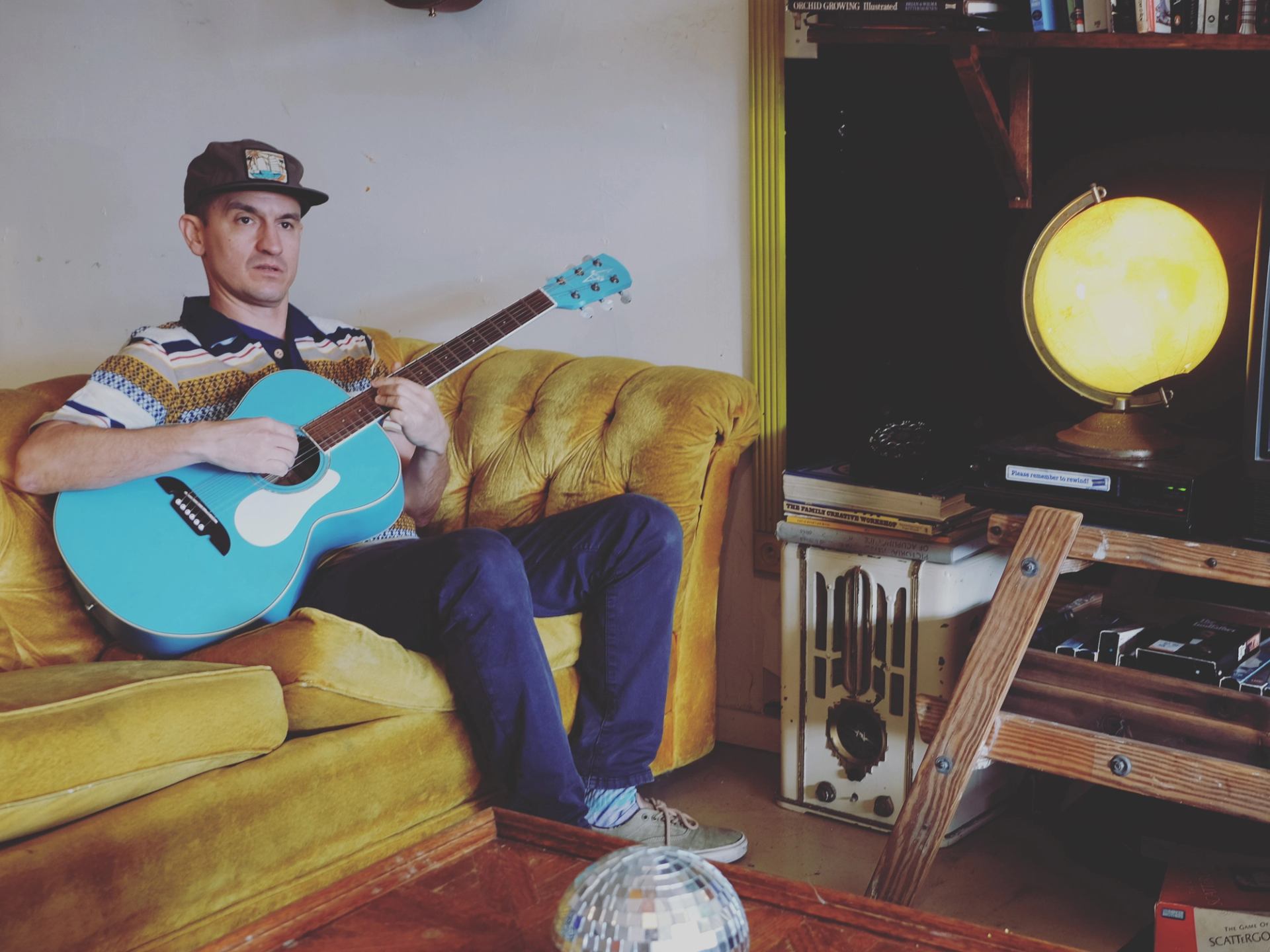 Wax plays guitar on a yellow couch in a cozy, eclectic room.