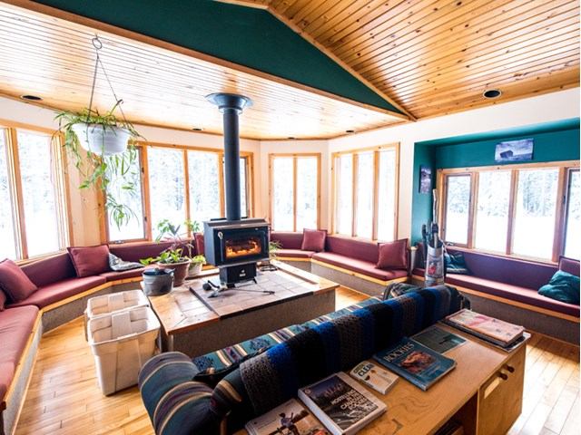 Warm cabin lounge with wood stove, red cushions, and sunlight streaming through large windows.