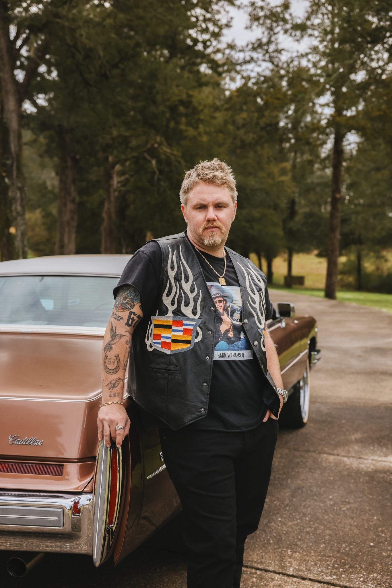 ERNEST wearing a leather vest leaning on a car with trees in the background.