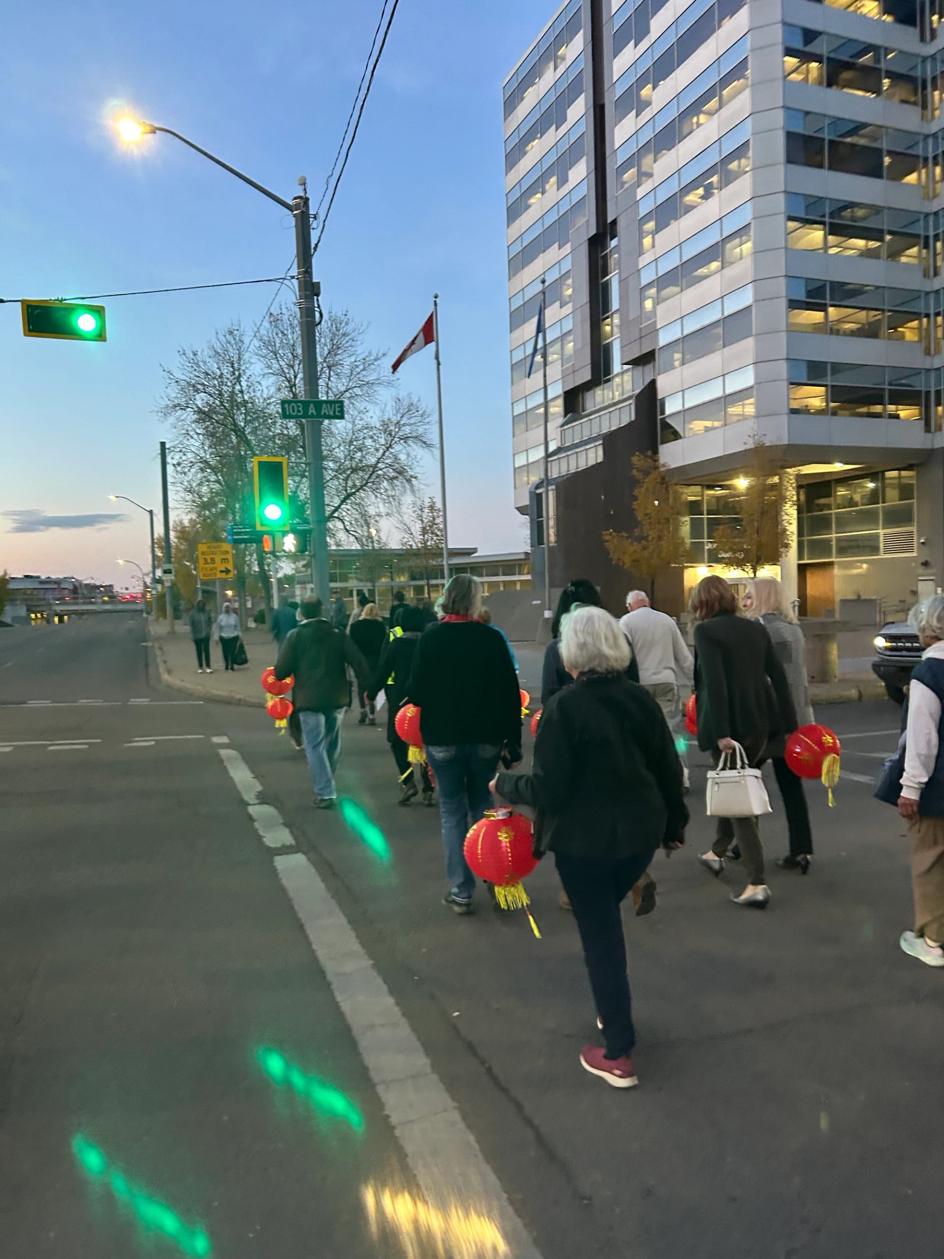 Pedestrians carrying red lanterns cross downtown street under green traffic lights.