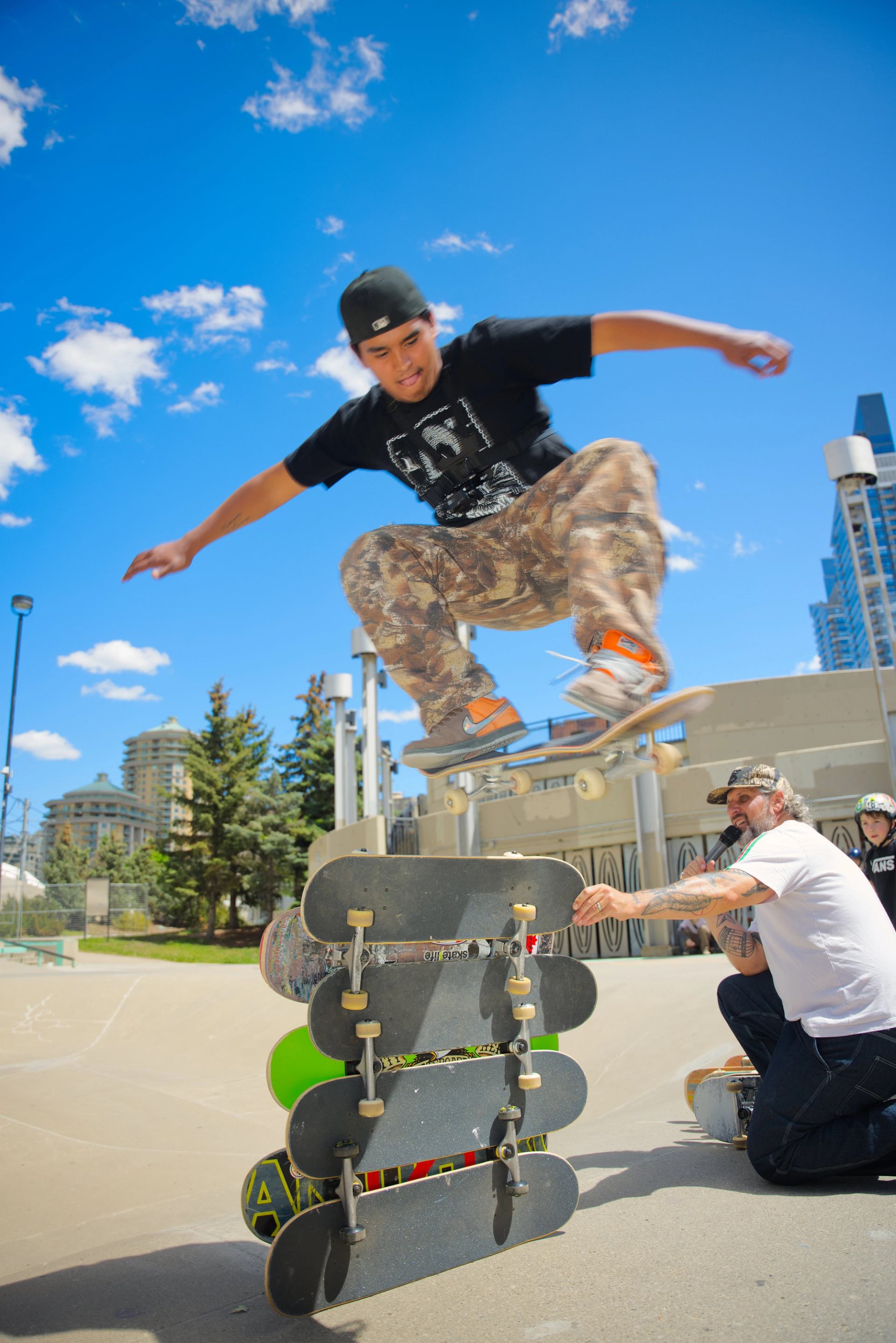 Skateboarder jumps over stacked skateboards at outdoor skate park under blue sky.