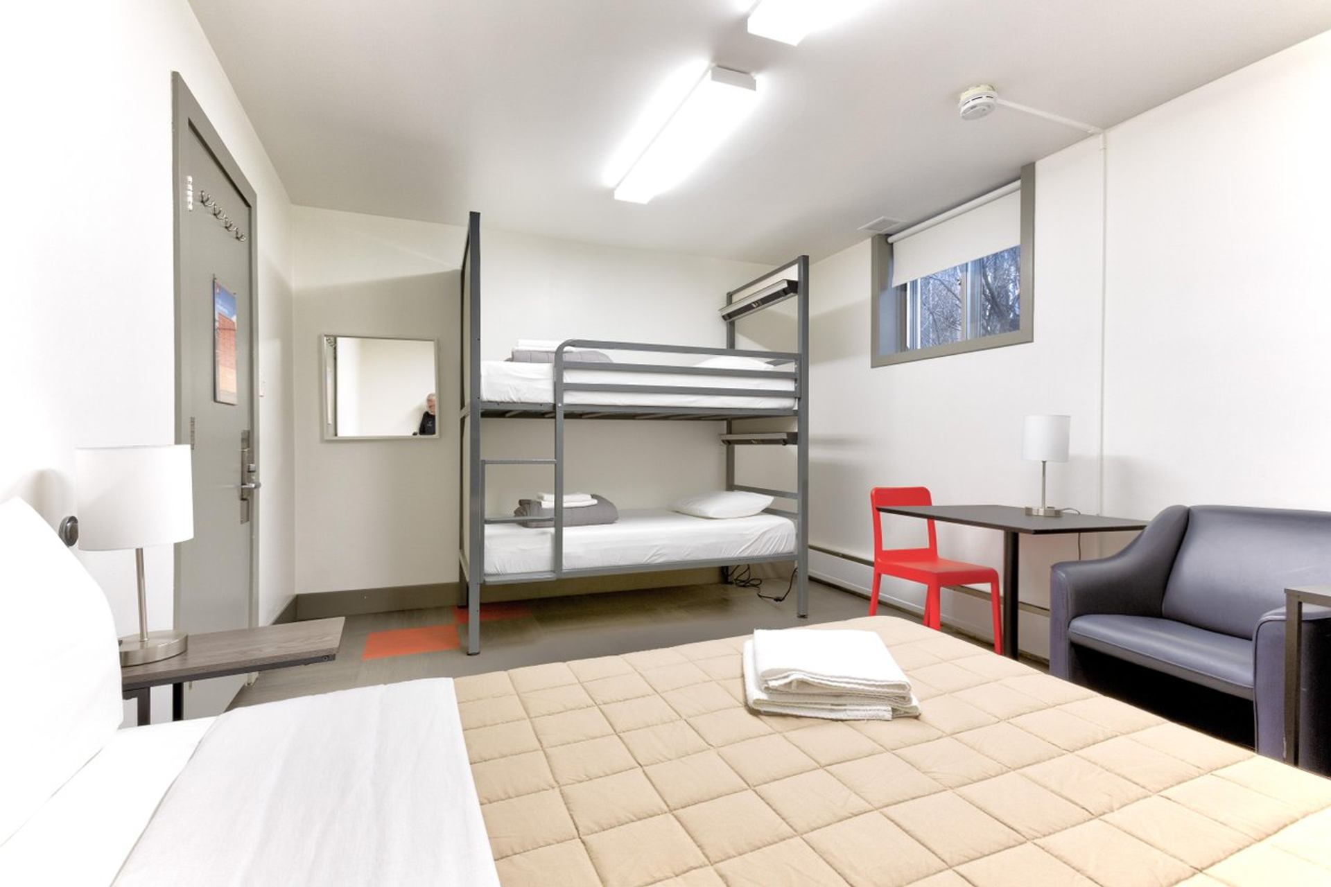 Family room with bunk bed, single bed, grey sofa, desk with red chair, and bright lighting.