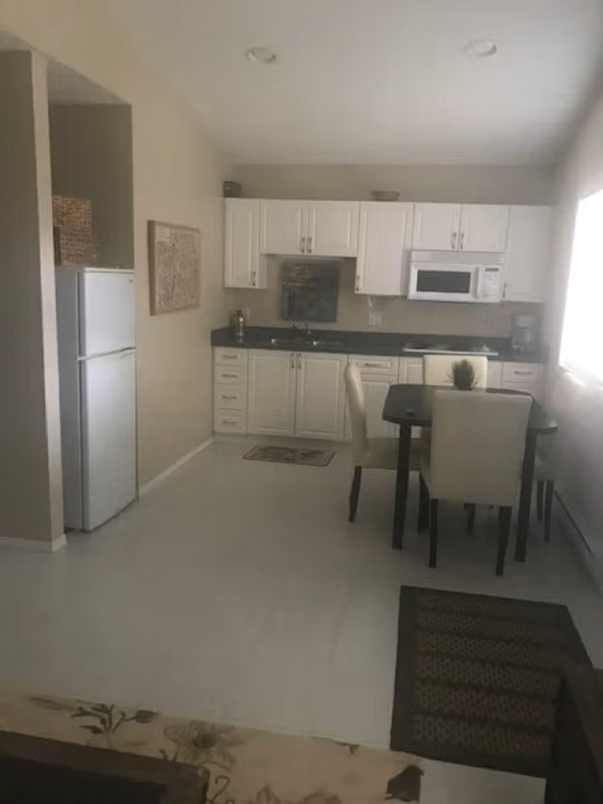 Small kitchen with white cabinets, dining table, chairs and refrigerator near window.