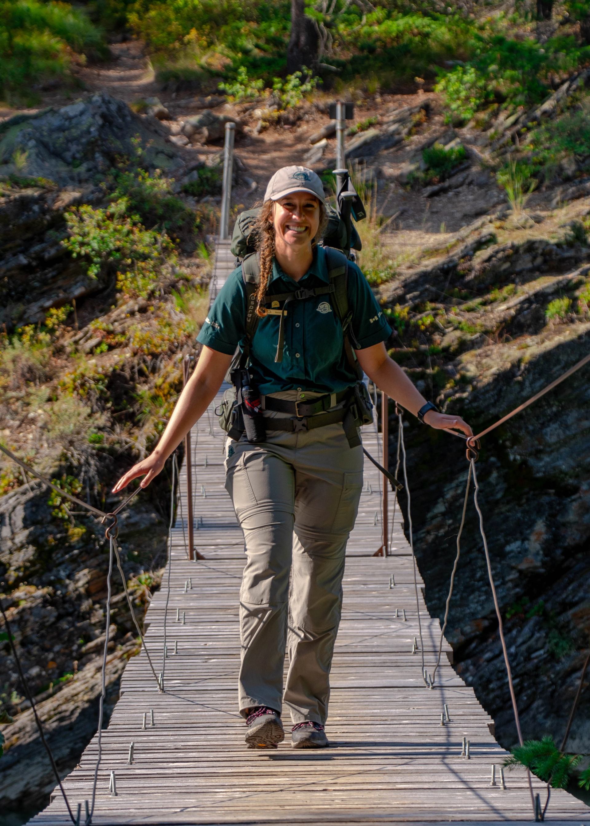 Hiker on wooden suspension bridge with cable railings, green hills and rocky trail behind.