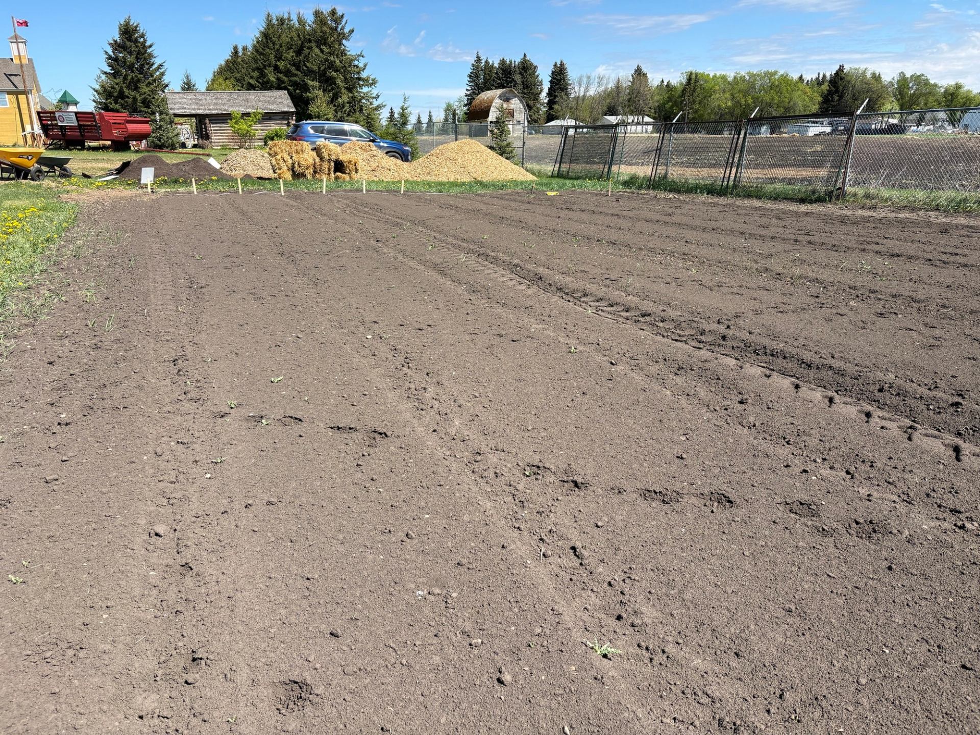 Freshly tilled garden plot beside chain-link fence, with buildings, hay bales and trees.