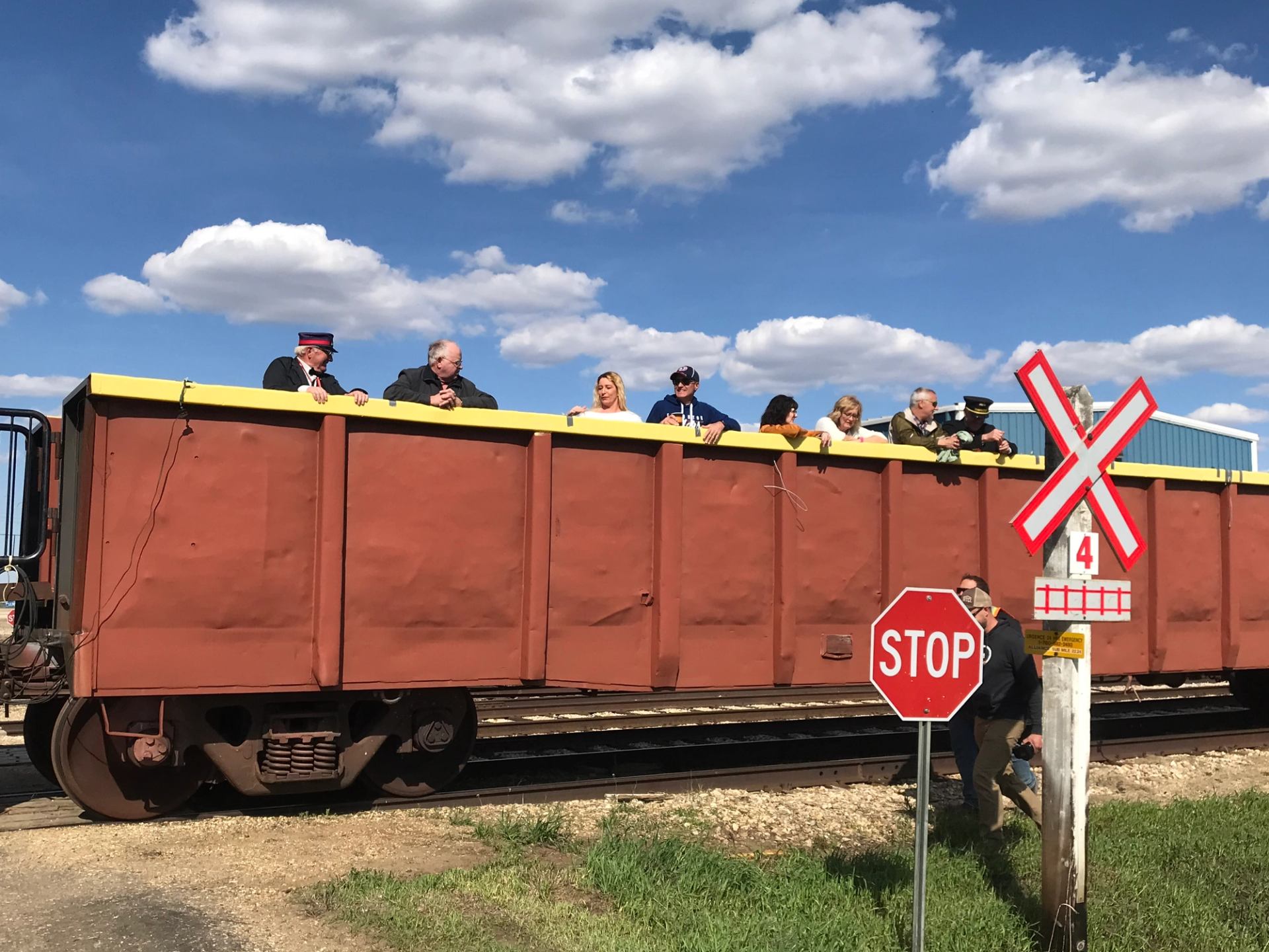Passengers riding in an open railcar beside a crossing sign under blue skies.
