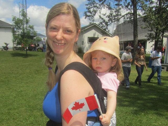 Adult carrying child in a carrier holds small Canadian flag at outdoor gathering.