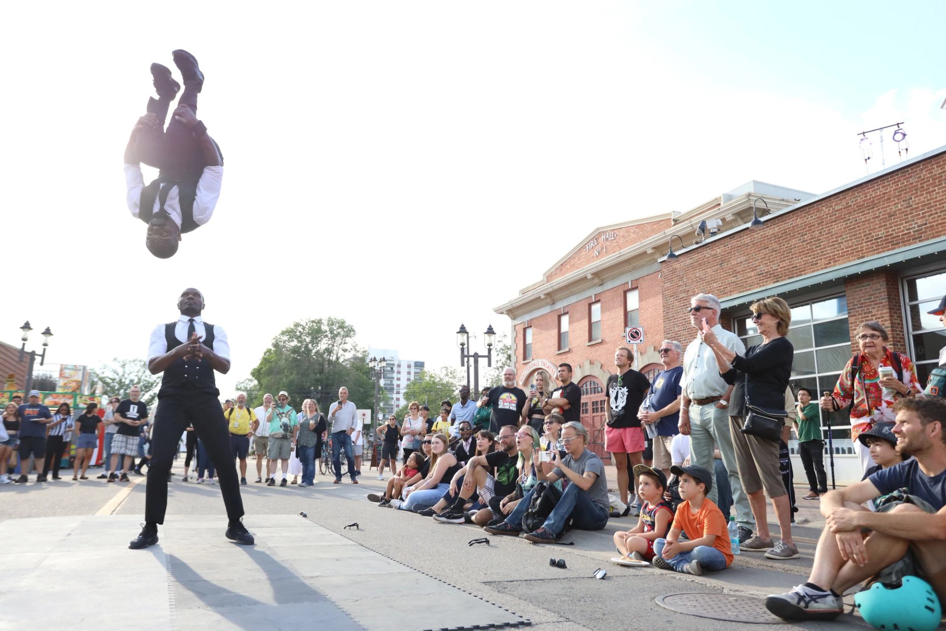 A performer in a vest and tie does a mid-air backflip over another performer as an outdoor audience watches.