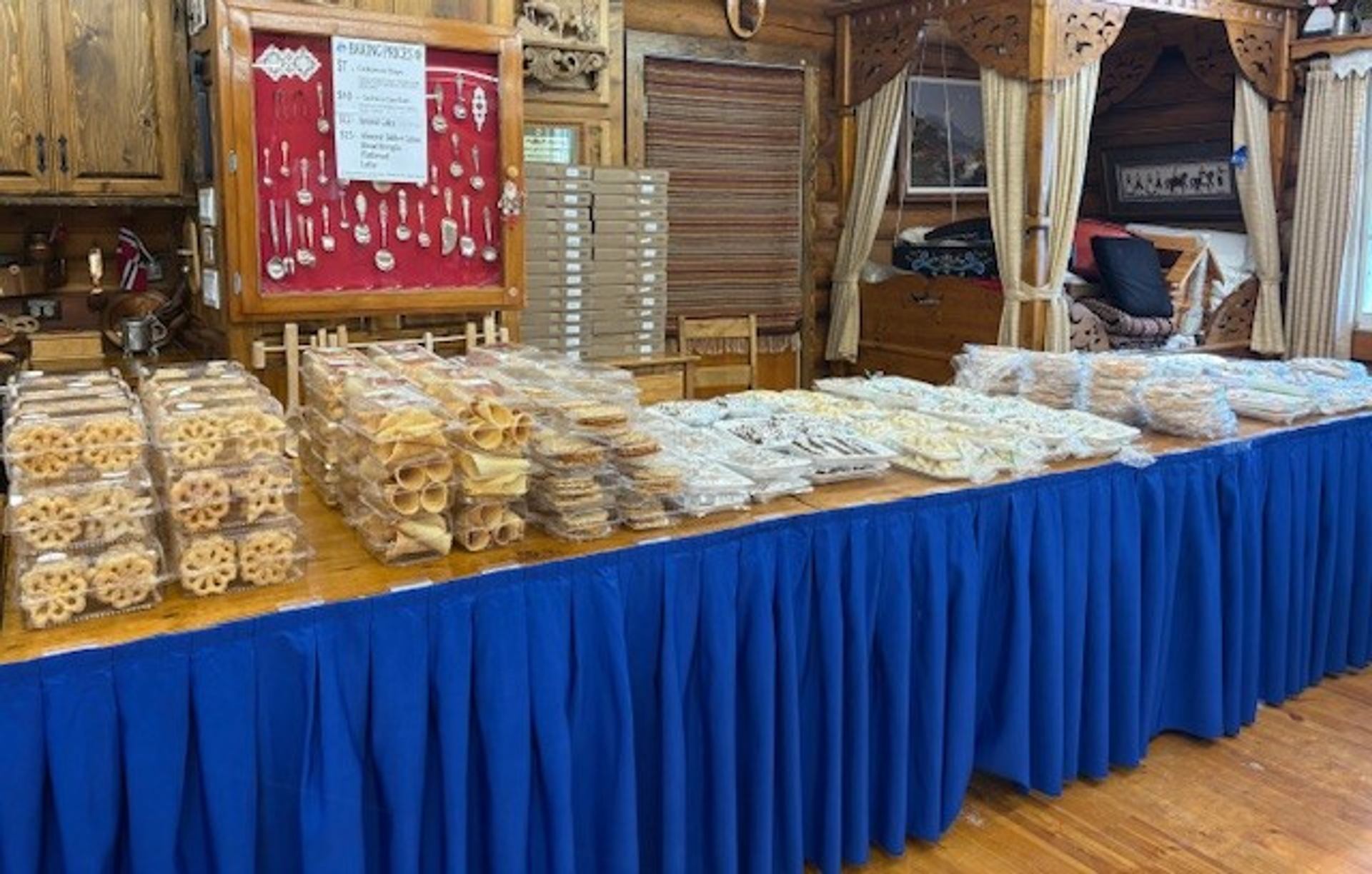 A long table draped in blue, laden with numerous pre-packaged baked goods, in a rustic wooden room.