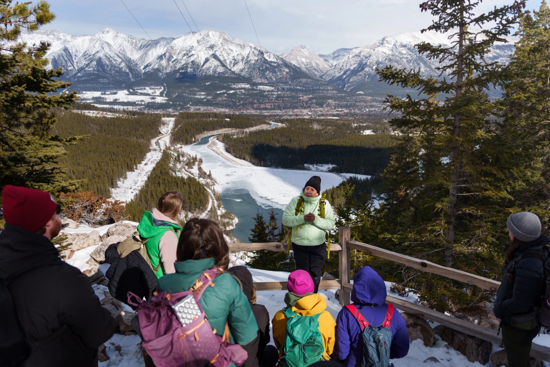 Group of hikers listening to guide overlooking snowy river valley and mountain range.