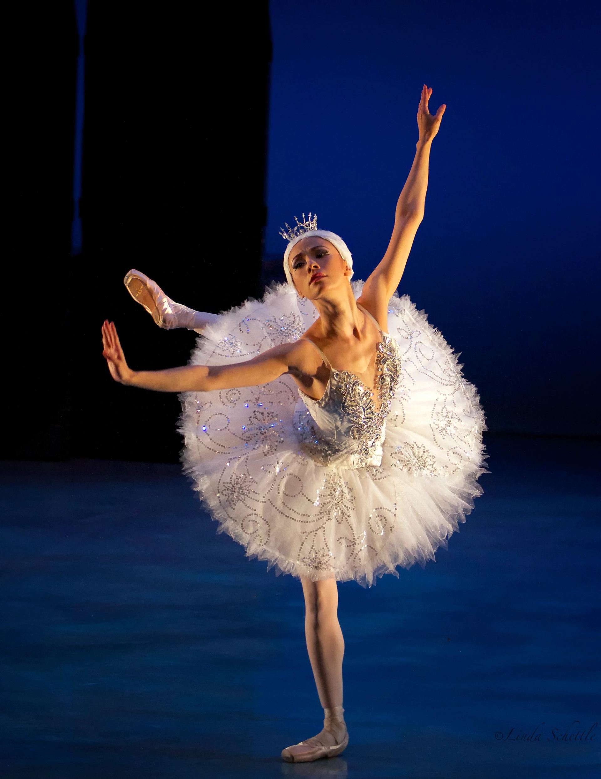 Ballet dancer in white tutu and tiara balances en pointe on blue-lit stage.