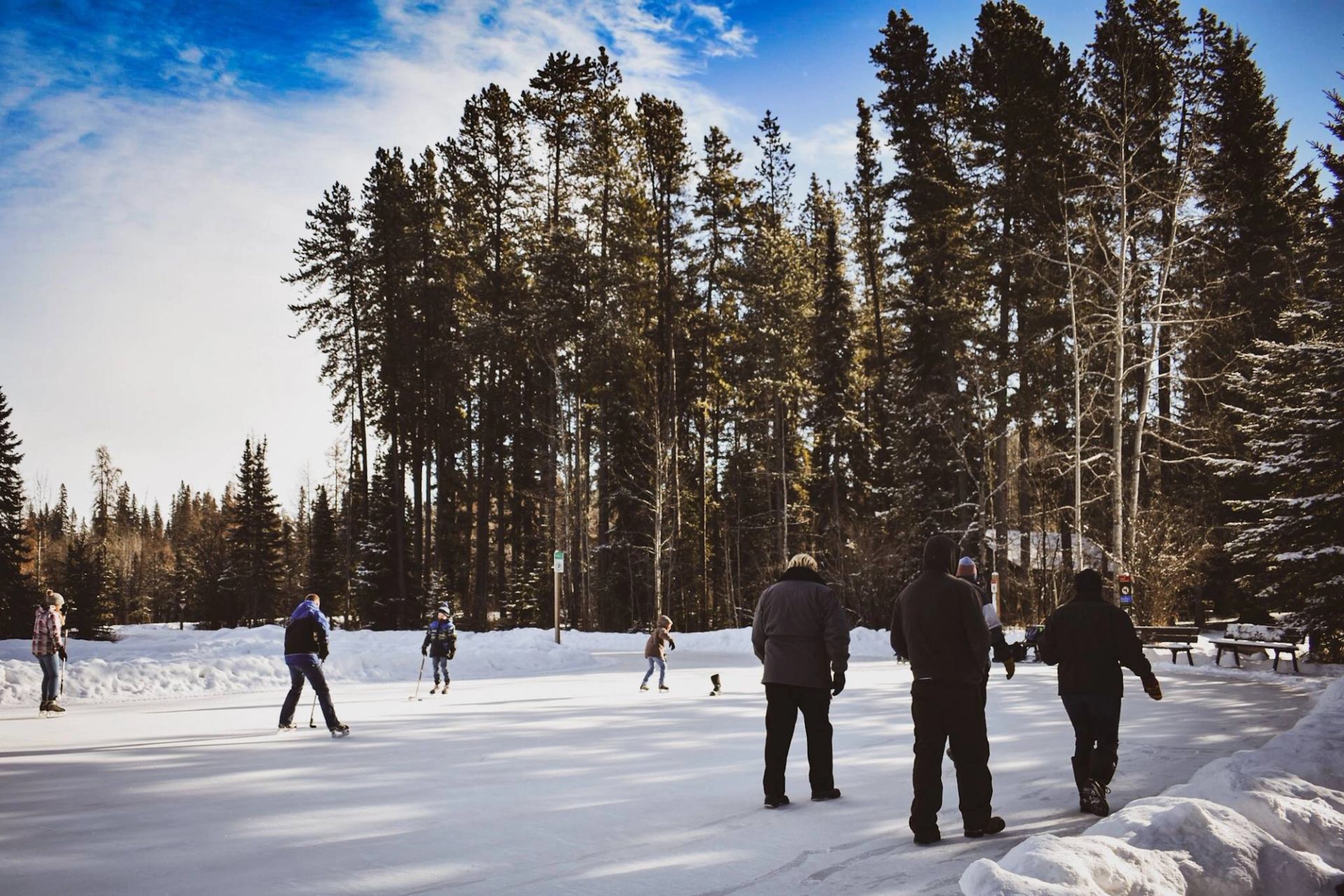 People ice skating on frozen rink surrounded by snow and evergreen trees.