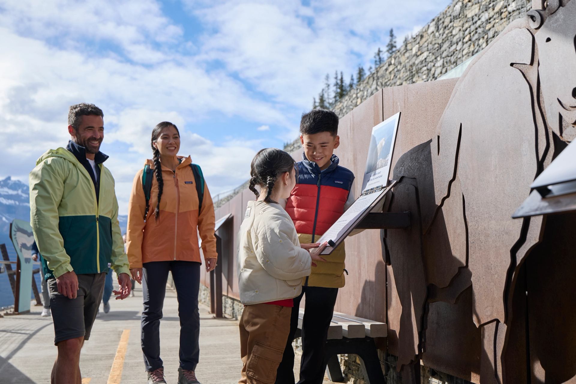 Visitors in jackets read an outdoor interpretive sign beside a metal animal sculpture.