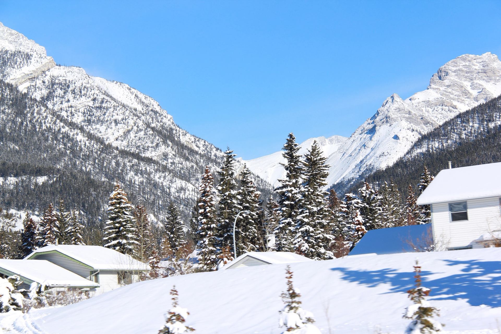 Snow-covered houses and evergreen trees below tall, rugged mountain peaks under clear blue sky..