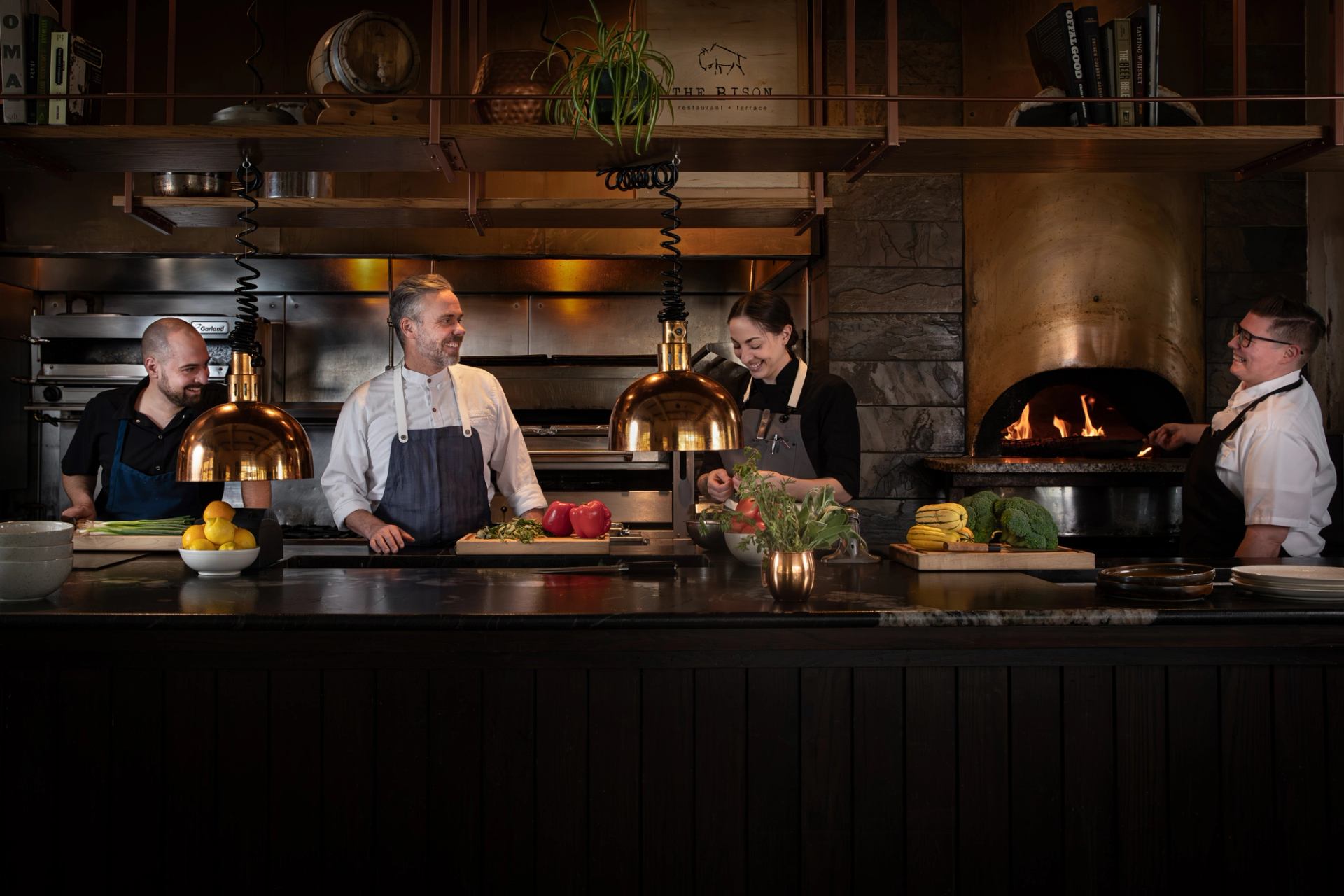 Four smiling chefs in a professional kitchen, preparing food with fresh ingredients near a wood-fired oven.