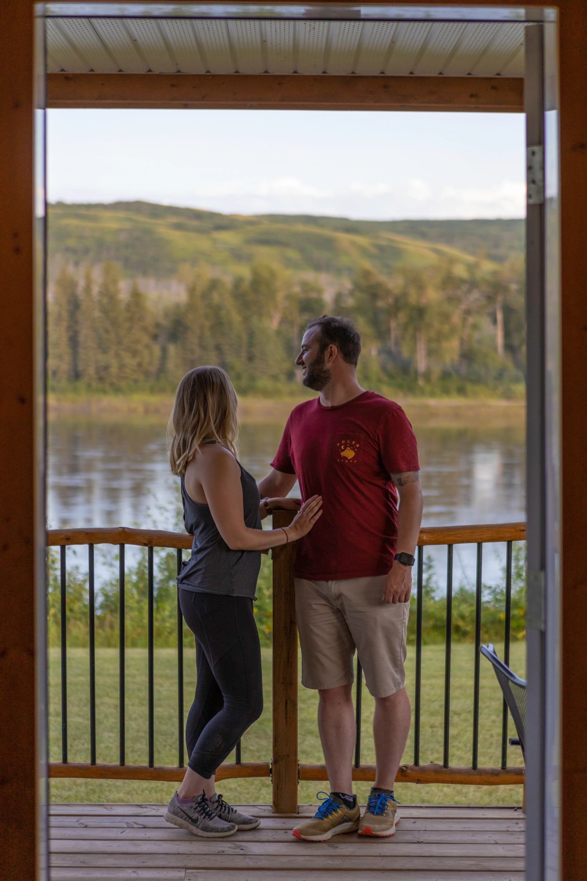 Two people standing on a wooden deck overlooking a calm river and green hills
