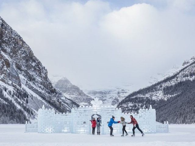 Skaters glide past an ice castle with snowy mountains in the background.