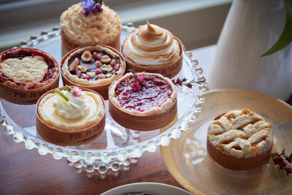 A glass cake stand holds six mini pies and tarts, including berry, chocolate, and meringue, plus a lattice-top pie on a gold plate.