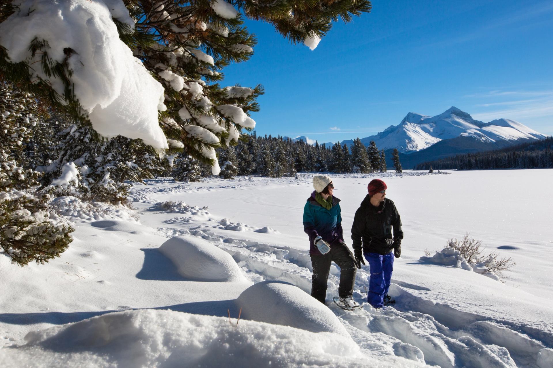Two people walking on snowy trail with mountains and trees in background