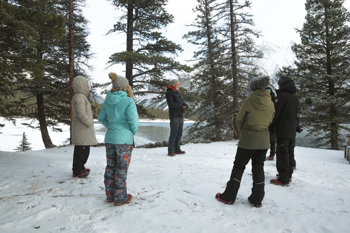 Group standing on snow in a forest clearing, dressed in winter gear near a frozen lake.