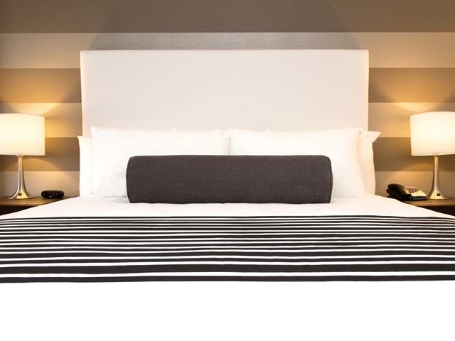 Modern hotel bed with striped runner, white linens, and bedside lamps.