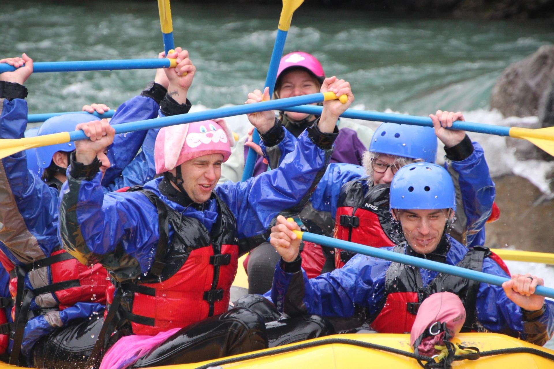 Group of rafters in helmets and lifejackets raising paddles on fast-moving river.