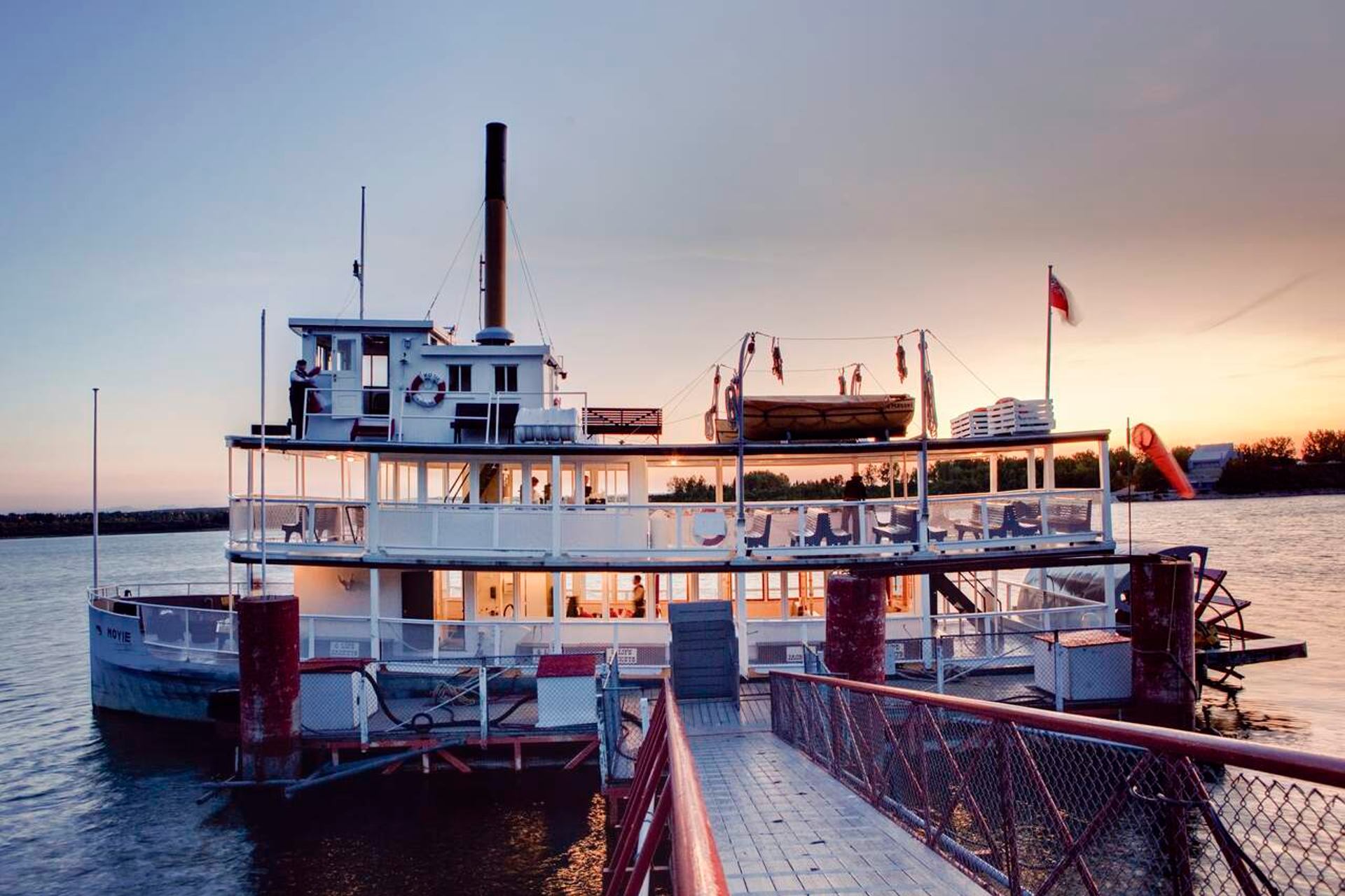 River paddlewheel boat docked at pier, lit interior, calm water and distant shoreline.