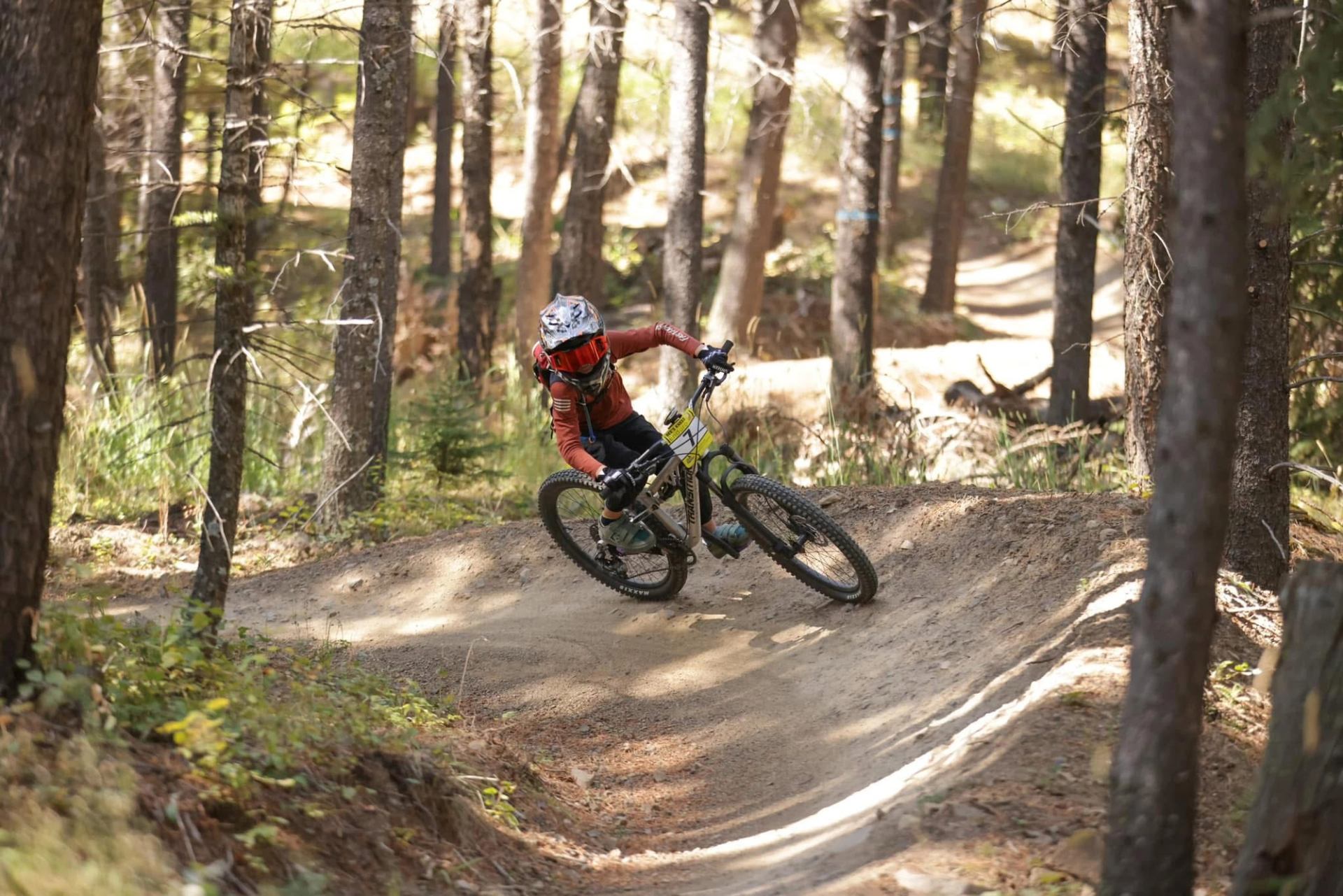 Mountain biker in red jersey rides dirt trail through forest trees.