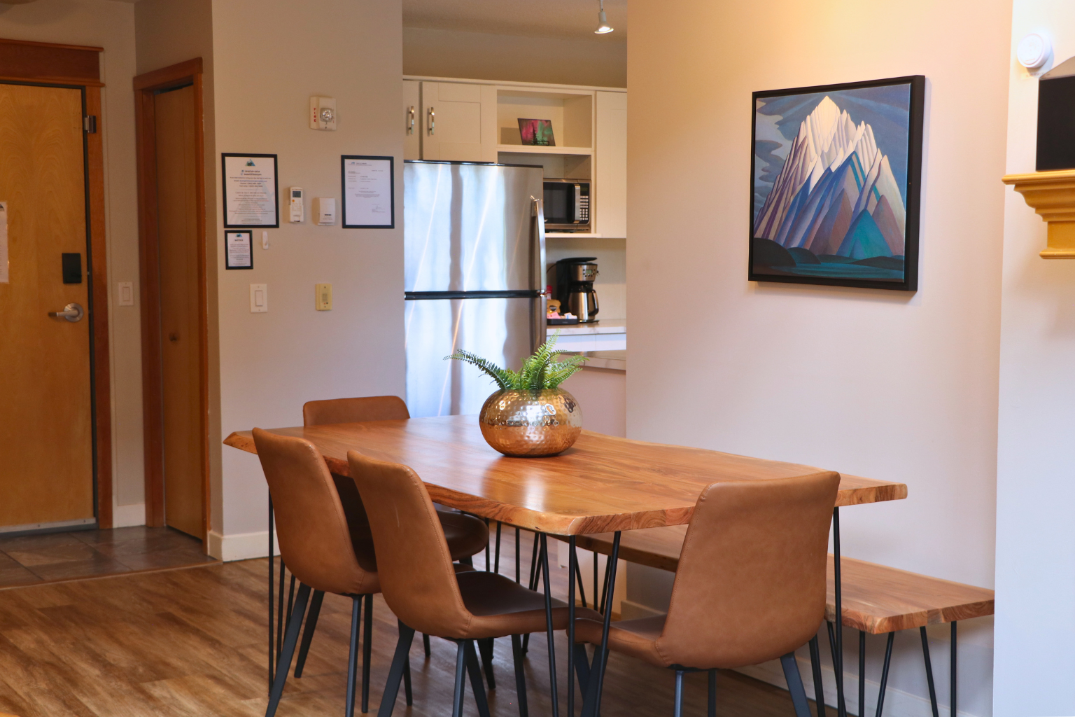 Wood dining table with brown chairs beside a kitchen and framed mountain artwork.