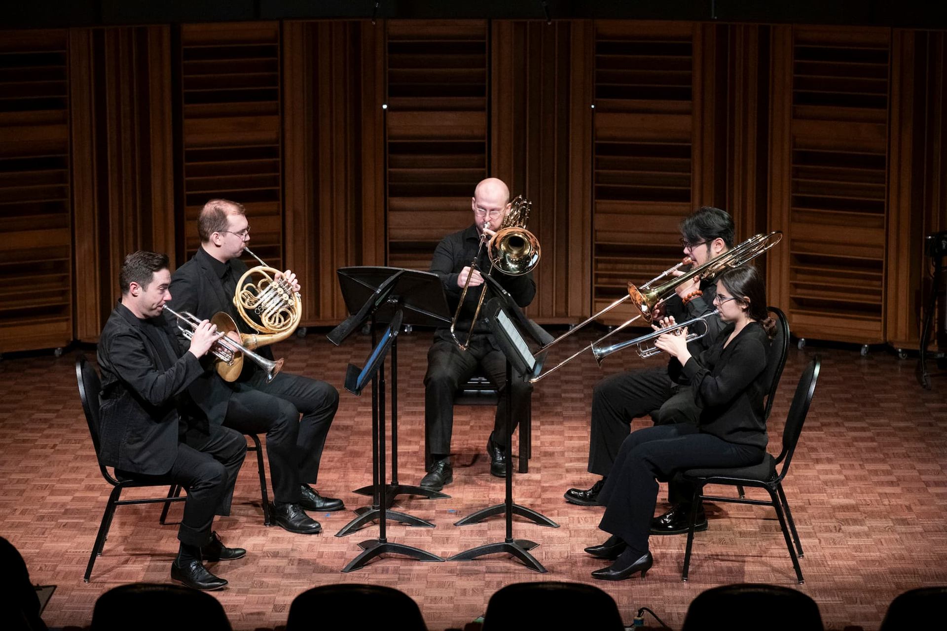 Brass quintet performs on stage with trumpets, trombones and French horn in concert hall.