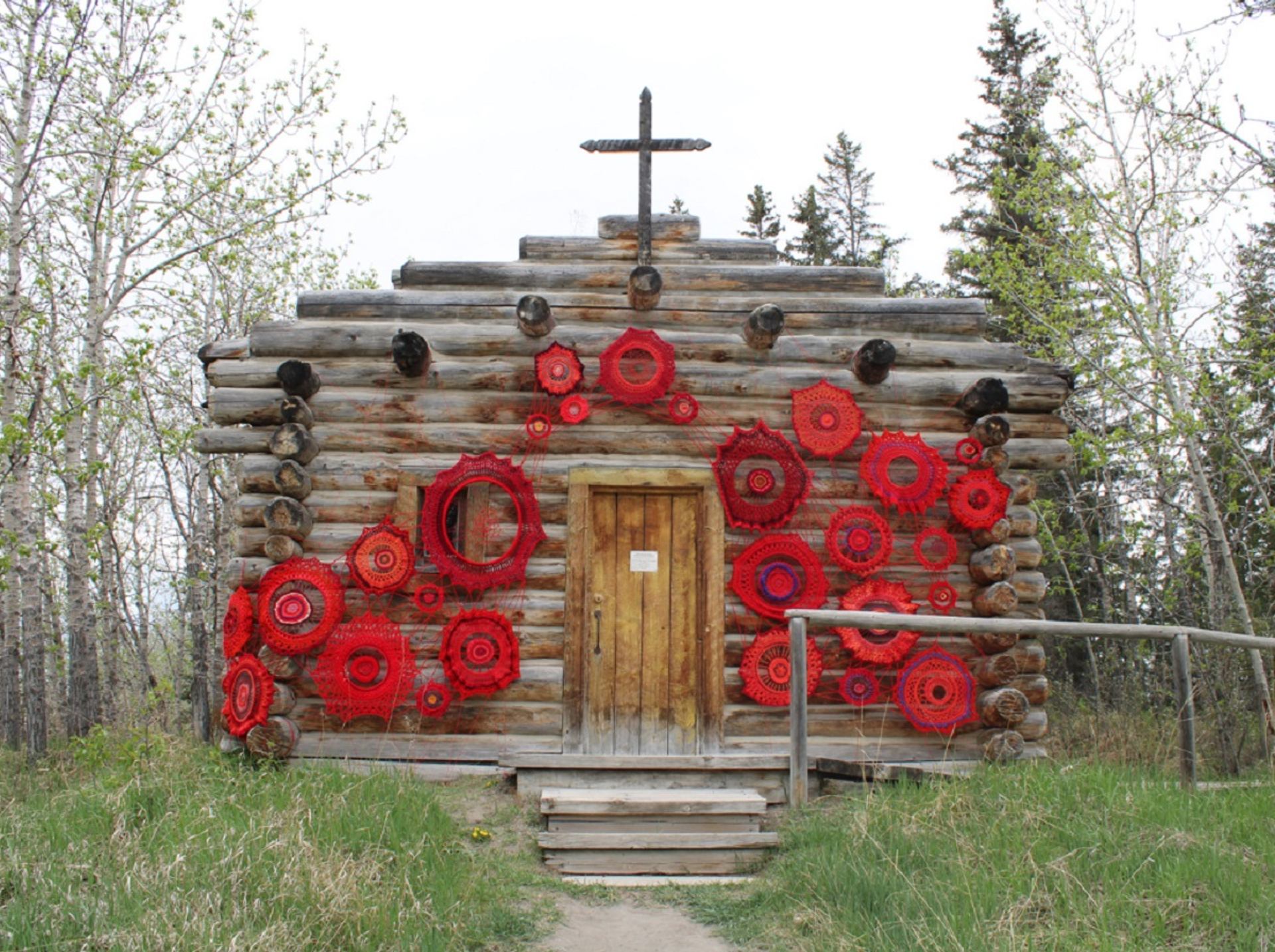 Log cabin with cross and red crocheted circles decorating exterior wall among trees.
