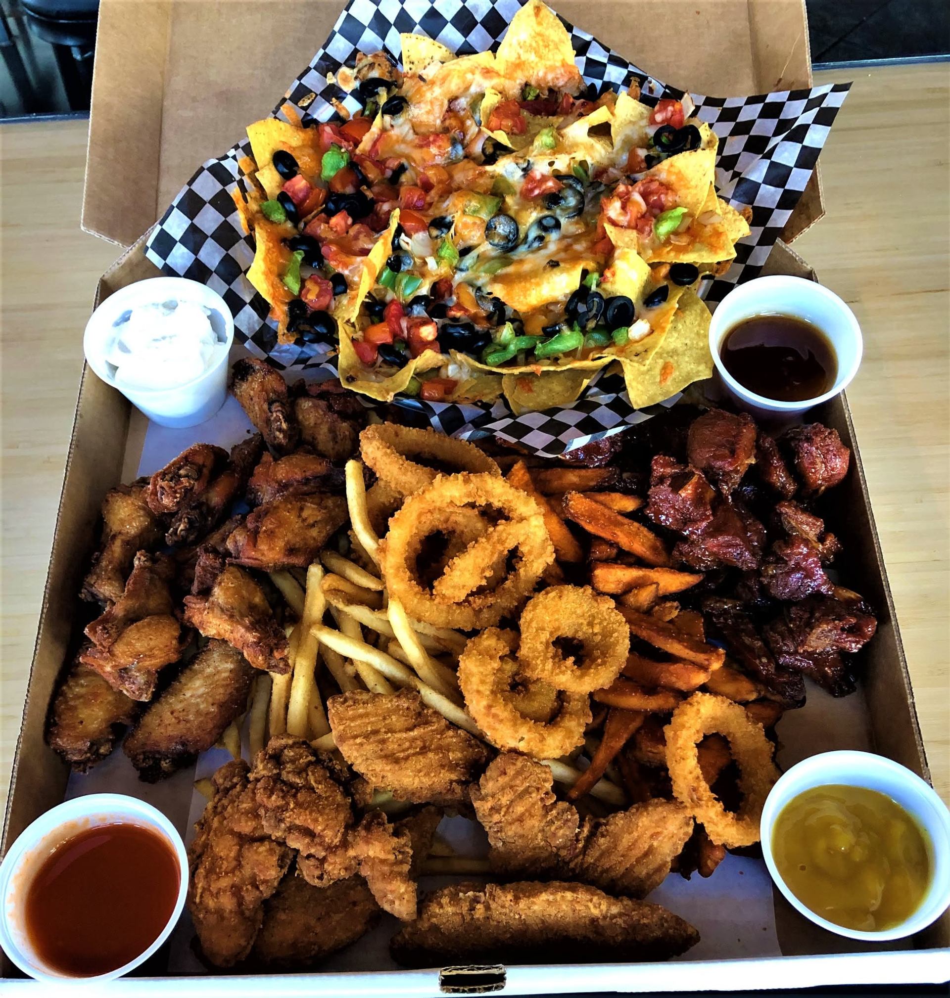 A platter features nachos, wings, onion rings, fries, chicken strips, and assorted dipping sauces.