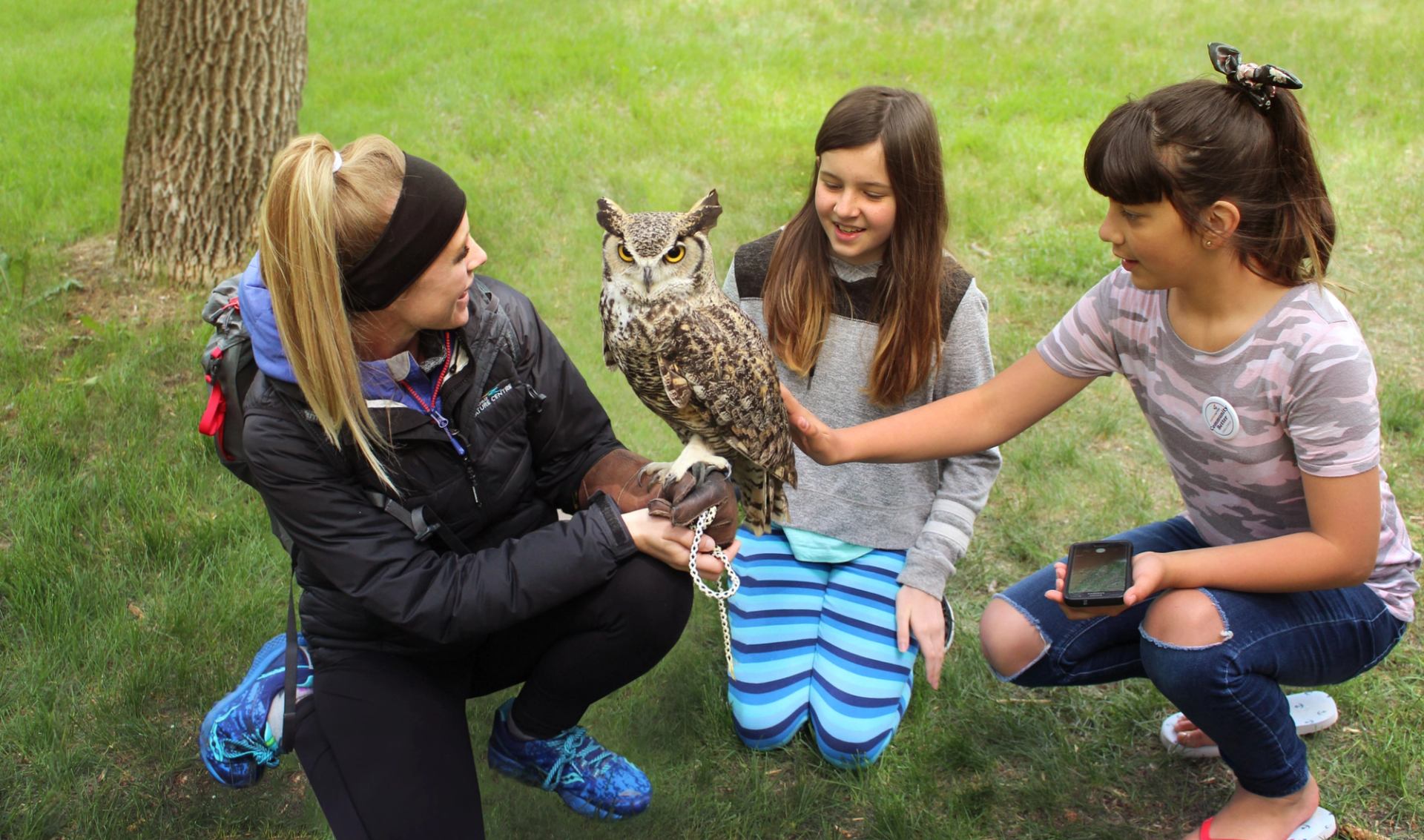Outdoor educator holding an owl while two children kneel nearby and gently touch it.