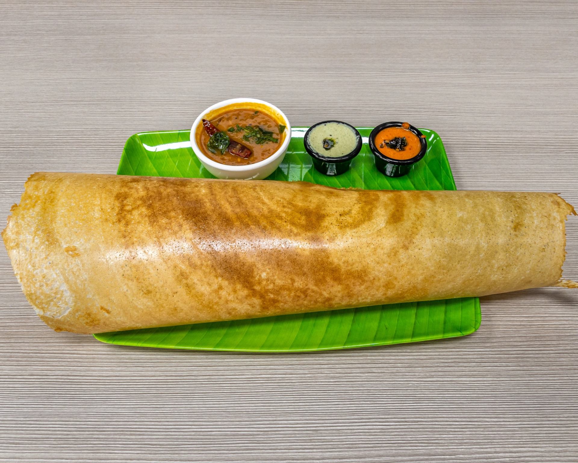 Large rolled dosa served with sambar and two chutneys on a green tray.