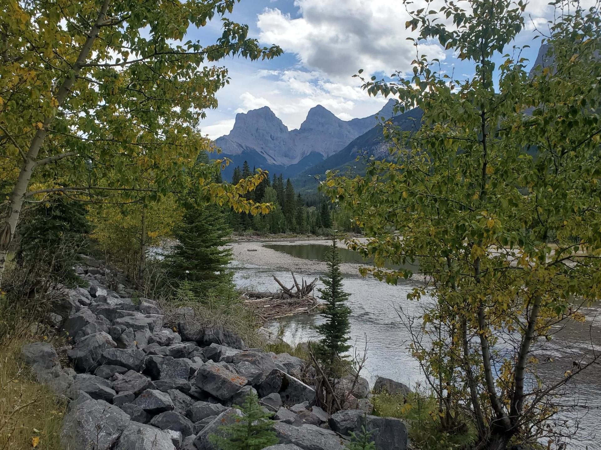 A flowing river bordered by rocks and trees with dramatic mountain peaks in the distance.