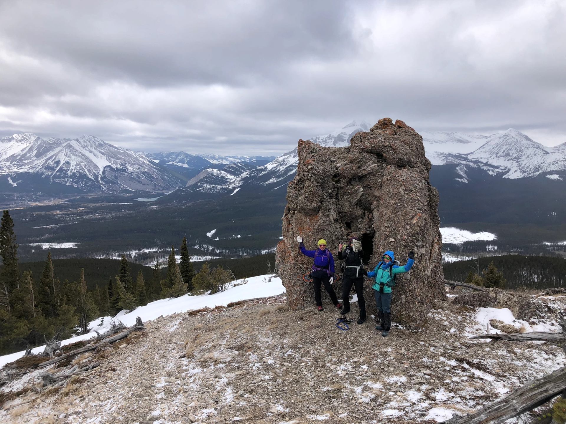 Hikers standing by a large rocky formation on a snowy mountain with panoramic peaks in the distance.