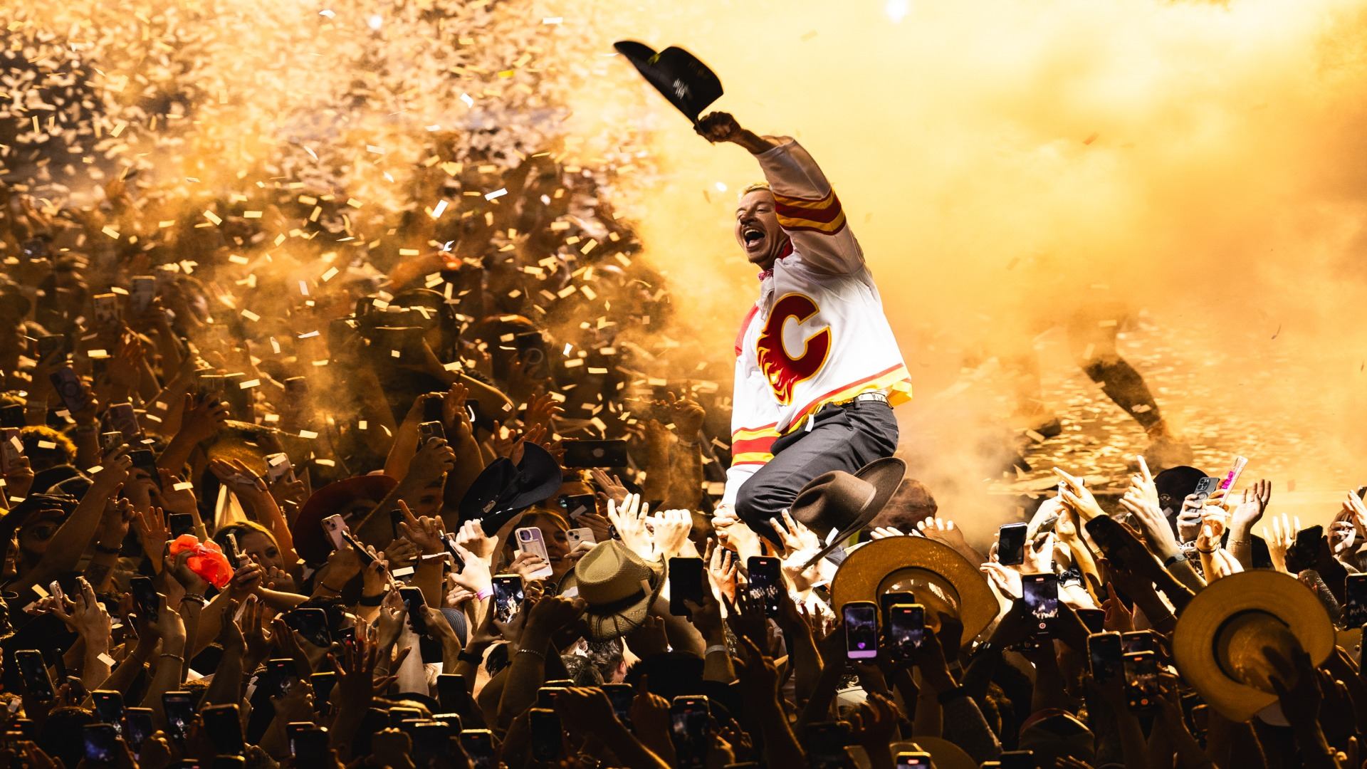 An man in a white Calgary jersey jumps with a hat in hand above a crowd, surrounded by golden confetti and orange smoke.