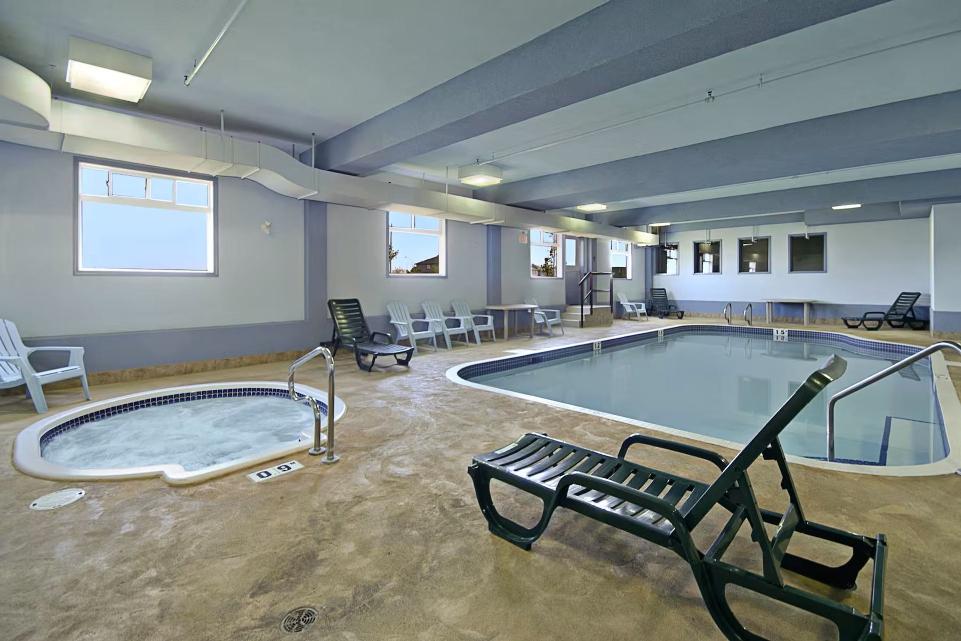 Indoor pool area with hot tub, lounge chairs, tables, windows, exposed beams, and calm, well‑kept surroundings.